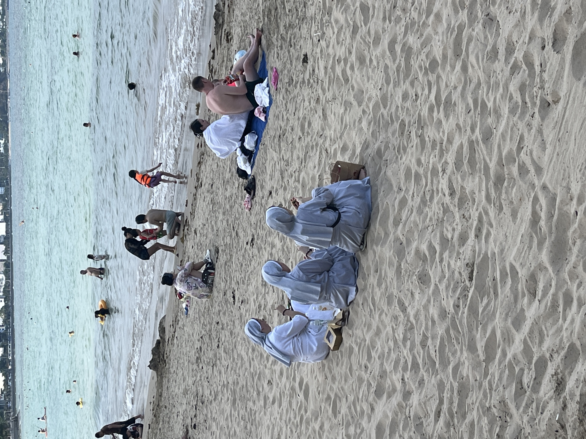 The image shows a beach scene in Bắc Nha Trang, Vietnam. Several people are in the water, swimming or wading. On the sand, there are groups of people: one group consists of a couple on a beach blanket and three people in white habits sitting together.