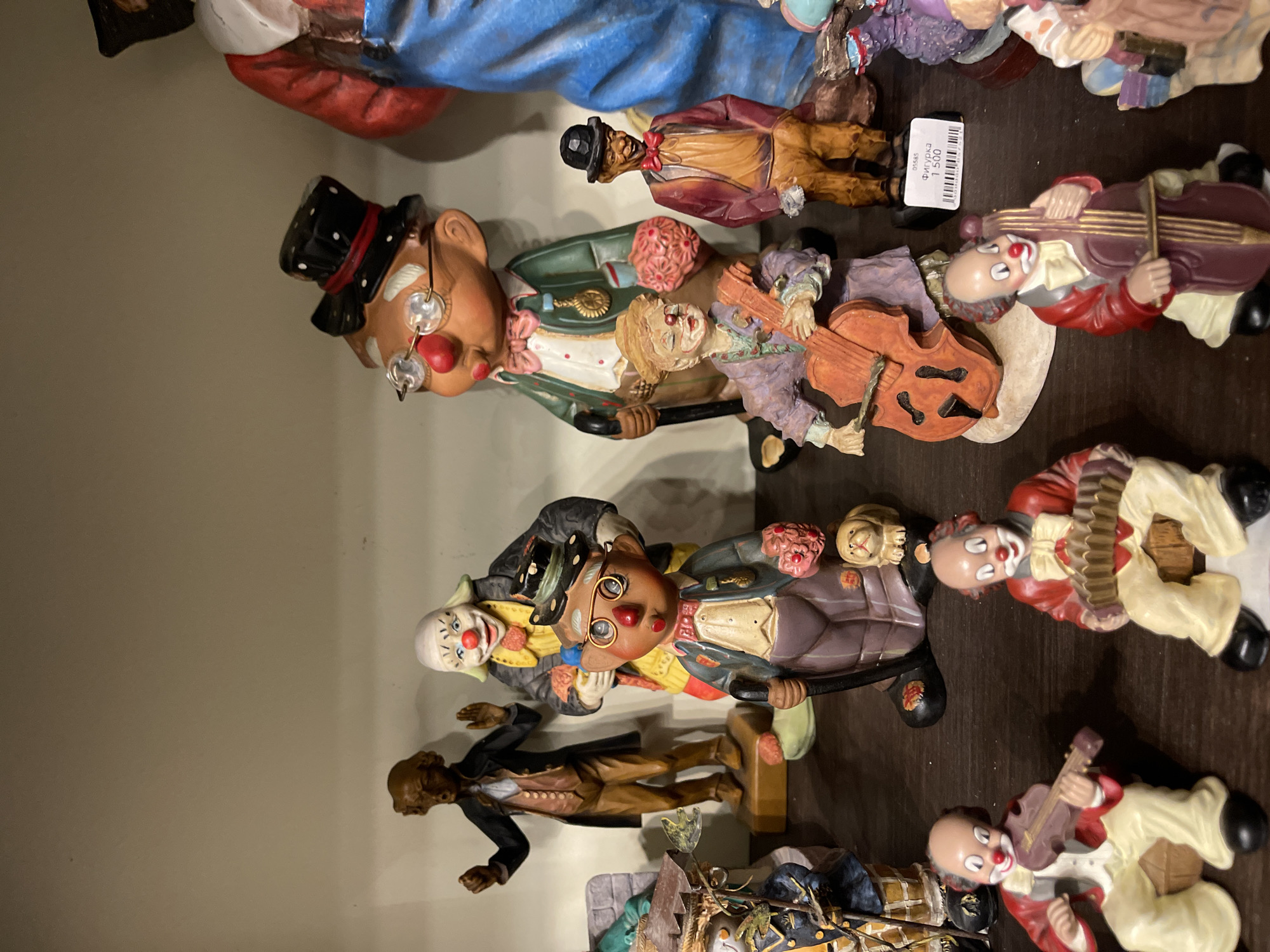The image displays a collection of vintage ceramic figurines, primarily featuring clowns, arranged on wooden shelves. The figurines are diverse in their attire and poses, with some playing musical instruments like violins and accordions, while others stand or hold props. In the background, a larger figure dressed as a historical gentleman is visible. A price tag is attached to one of the figurines. The overall setting appears to be a shop or a private collection. The location is identified as the Museum of Chernyakhovsk porcelain in Svetlogorsk, Russia, indicating it's a museum display.