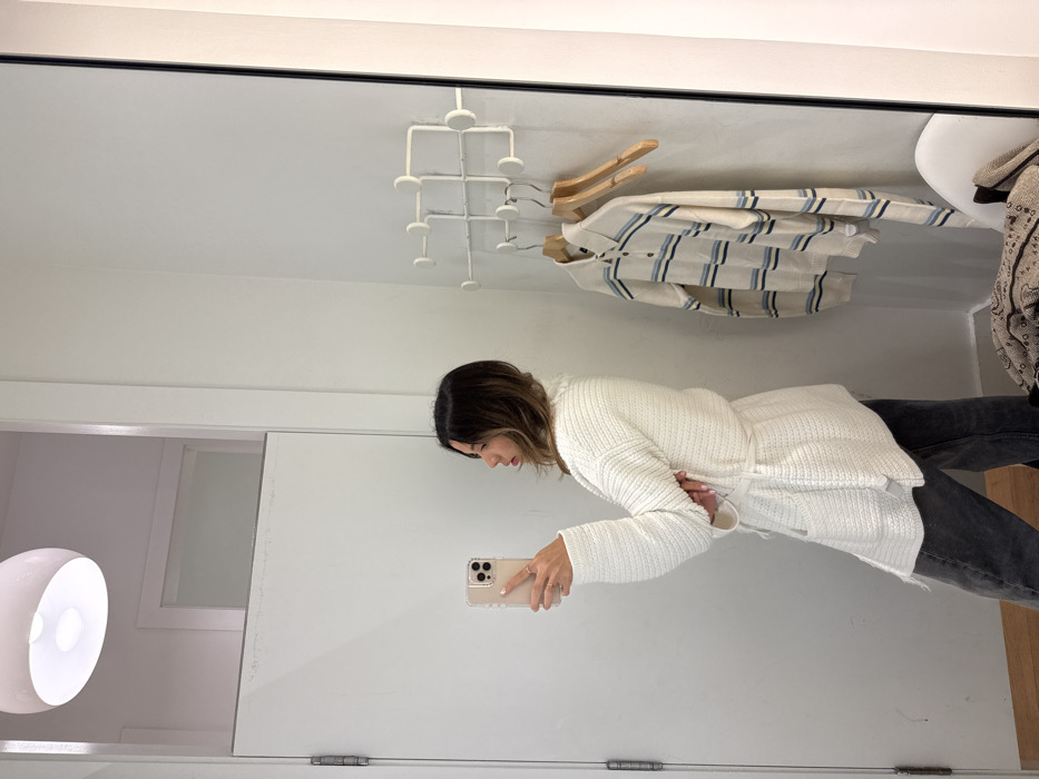 In a mirror selfie, a woman wearing a white knit sweater and dark jeans stands in what appears to be a dressing room. A white light fixture is visible on the left. Clothes hang from a rack at the top, including a blue-striped shirt on a hanger. A white organizational structure is also present on the ceiling. The mirror frame is visible at the top and left edges.