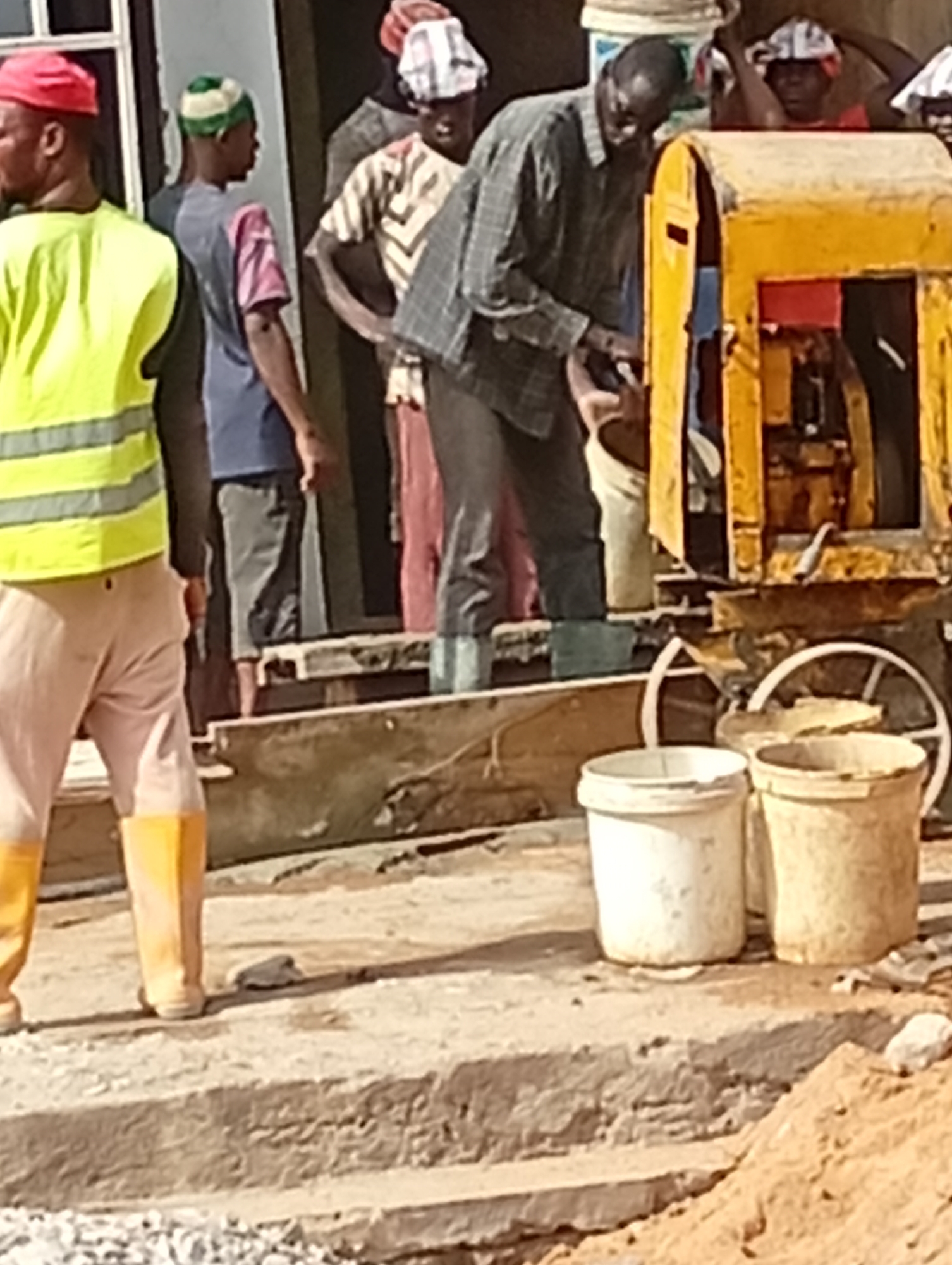 The scene depicts construction work in Maiburiji, Nigeria. A group of men is gathered around a yellow cement mixer, with one man actively feeding material into the machine using a white bucket. Two additional white buckets sit on the ground nearby. Other men are standing around observing the work. One man is wearing a yellow reflective vest and boots. The environment appears to be an outdoor construction site with sand and gravel visible.