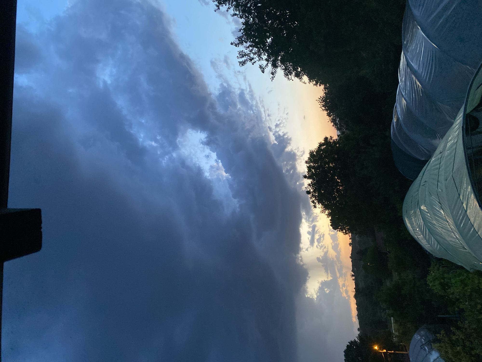 The image depicts a cloudy sky at dusk, with dark storm clouds dominating the left side of the frame and lighter, sunset-tinged clouds visible towards the right. The horizon line is partially obscured by trees and possibly a distant hill, suggesting a rural or semi-rural location. A partially visible dark structure on the left side suggests an object in the foreground. On the right, there are light-colored rounded structures, possibly greenhouses, and the faint lights suggesting a settlement. It is not possible to confidently confirm the location as Konstantinovo, Bulgaria. There are no people visible in the image.