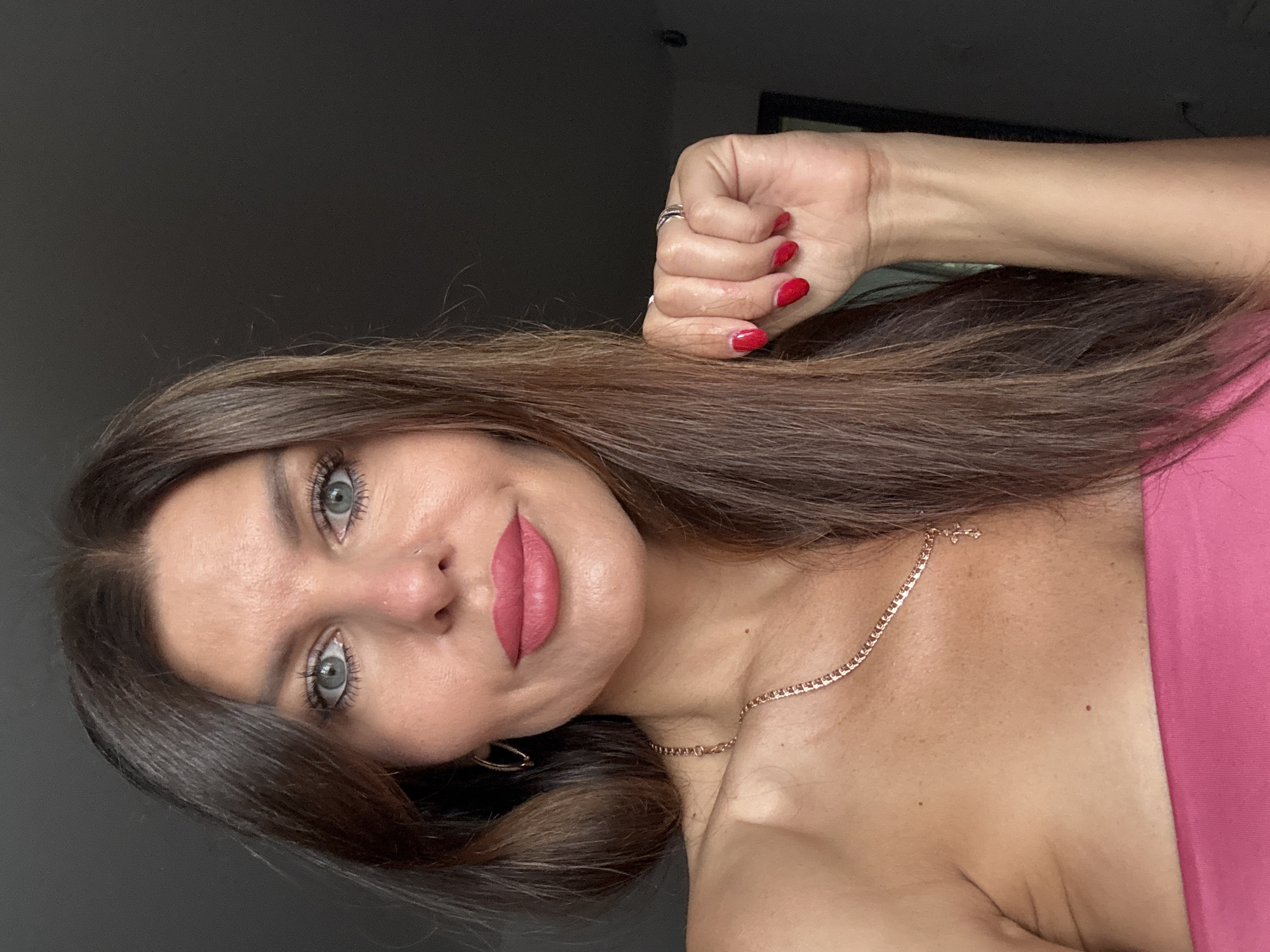 A medium close-up shot captures a woman in her 40s with long brown hair, fair skin, and blue eyes. She is wearing a strapless pink top and a gold necklace with a cross pendant. Her lips are painted with a bright pink lipstick, and her fingernails are painted red. She has a gold hoop earring in her left ear and two rings on her right hand. Her right hand is raised and held near her face. The background is dark and out of focus, with a glimpse of a mirror on the right side of the frame. The image is taken indoors. The location is not specified in the image.