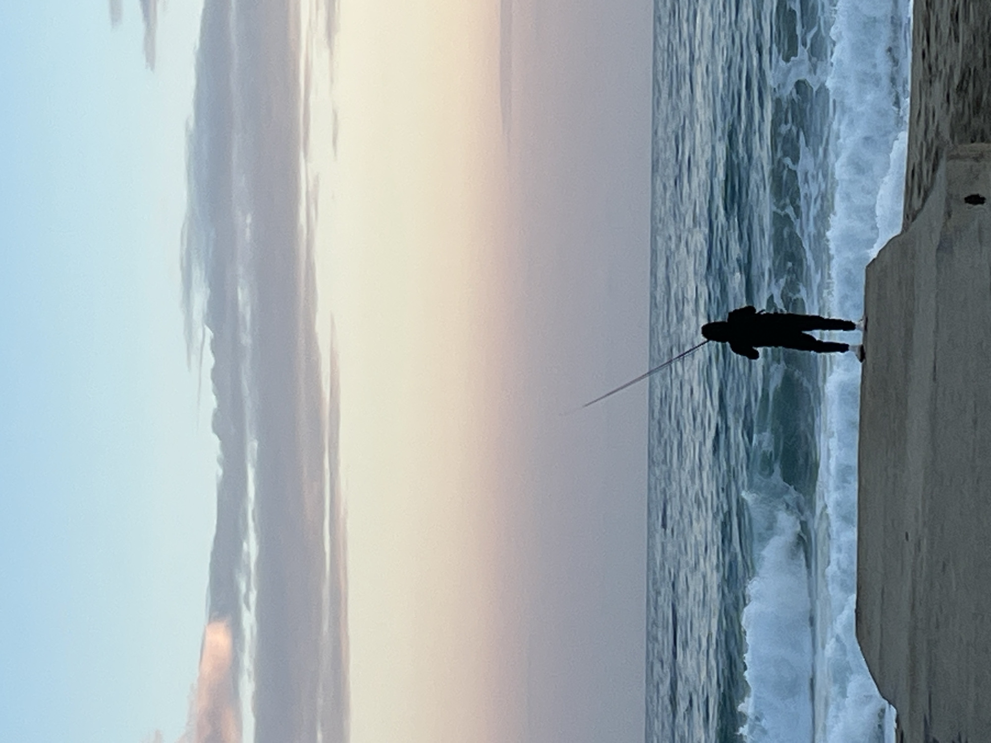 A silhouetted person fishes from a concrete embankment into a turbulent sea. The sky blends blue and soft pink hues, with elongated clouds parallel to the water. It is located in Alanya, Turkey.