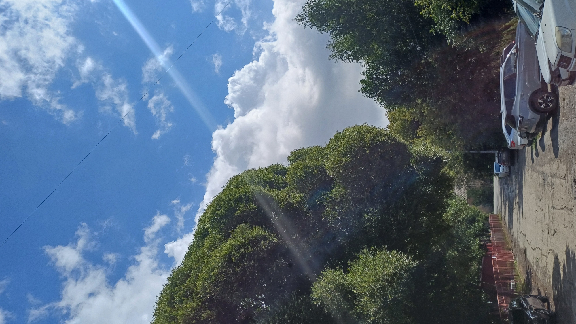 The image depicts a street in Izhevsk, Russia, on a bright, partly cloudy day. Dense green foliage from large trees lines both sides of a paved road, which shows signs of wear and tear with visible cracks. Several cars are parked along the right side of the street. The sky above is a vibrant blue, with fluffy white clouds scattered across it, and a diagonal ray of sunlight cuts across the upper right portion of the frame. A thin power line runs diagonally across the sky. On the left side of the road, a red metal fence encloses an area. The scene suggests a residential or suburban area.