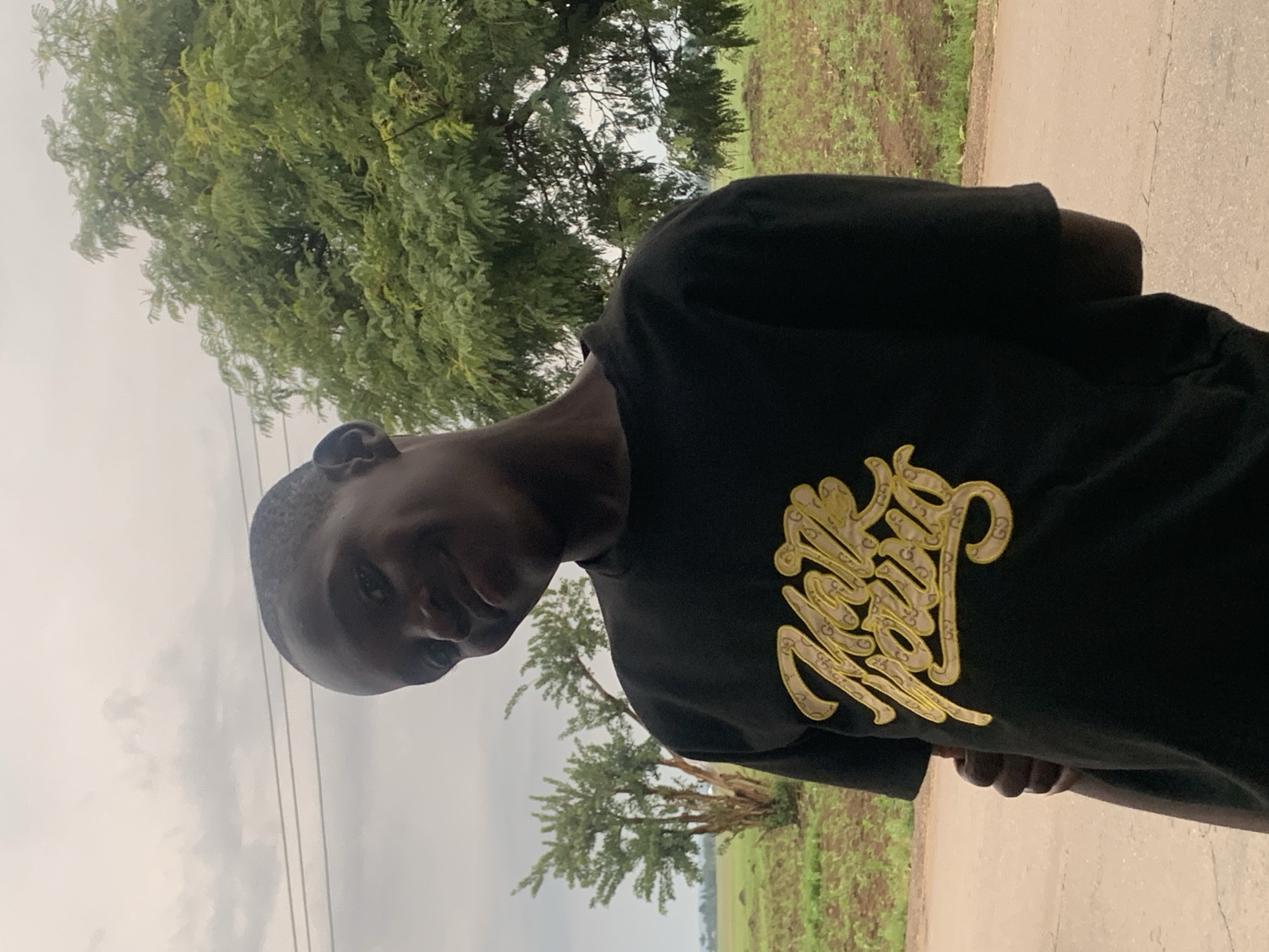 In Agban, Nigeria, a young Black male stands outdoors, head tilted slightly to his left and looking at the camera with a slight smile. He is wearing a black t-shirt with a gold, script-like logo on the front. The background consists of green foliage and a cloudy sky.