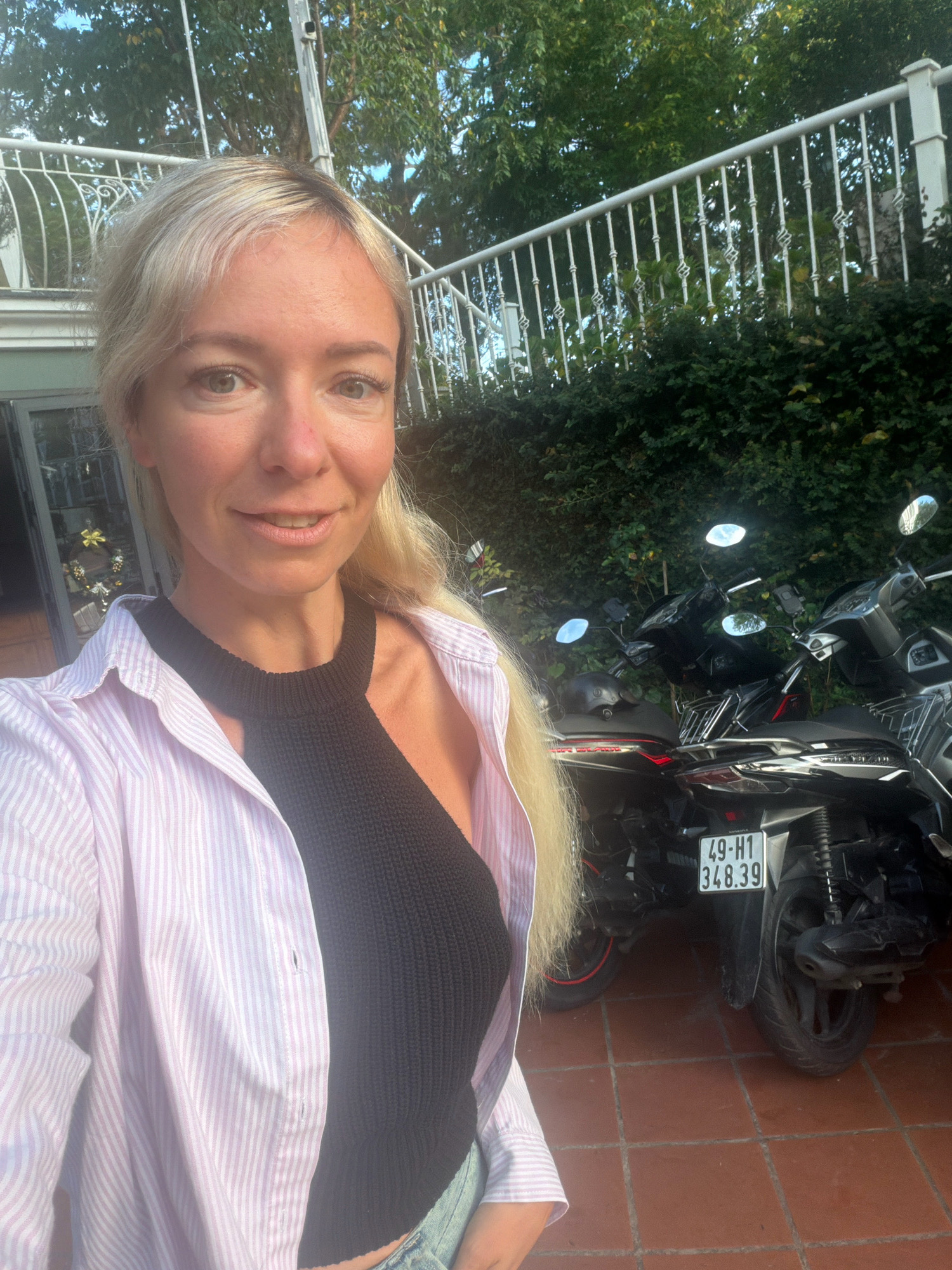 In Xuân Trường, Đà Lạt, Vietnam, a woman with blonde hair is taking a selfie. She is wearing a pink and white striped unbuttoned shirt over a black knitted top and blue jeans. Behind her, several motorcycles are parked on a terracotta tile floor. The background shows lush green foliage and a white railing. The license plate on one of the motorcycles reads "49-H1 348.39". The lighting suggests it is daytime, likely late afternoon due to the warm tones and shadows.