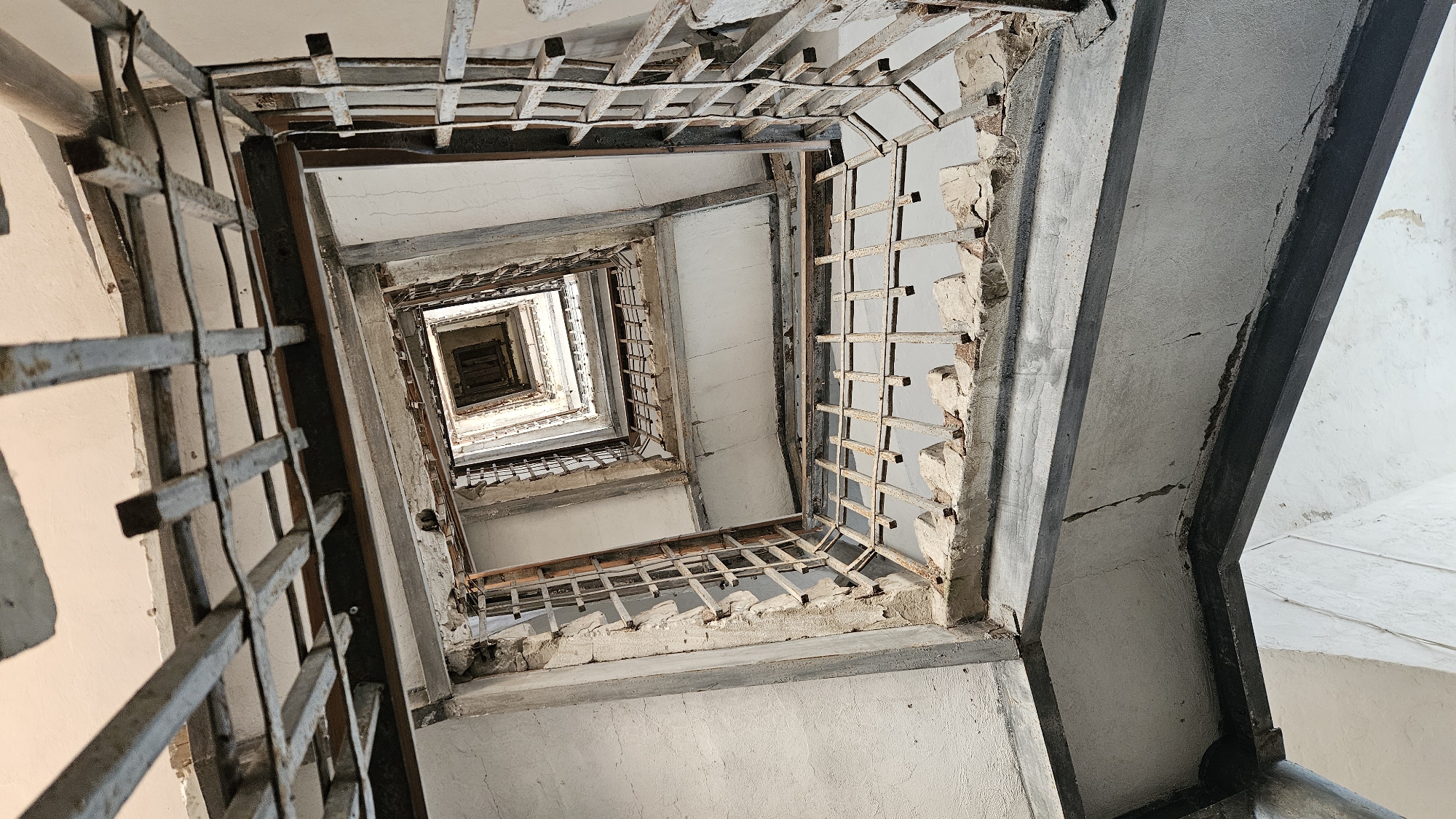 An upward perspective shot shows a decaying, square-shaped stairwell in an abandoned building. The concrete walls are stained and cracked, and the metal railings are rusted and partially broken, with visible damage to the concrete ledges supporting them. The stairwell ascends into a dark, recessed opening at the top, suggesting multiple floors. The environment appears derelict and neglected, with no signs of current human activity. This scene is not located in Suzdal, Russia.