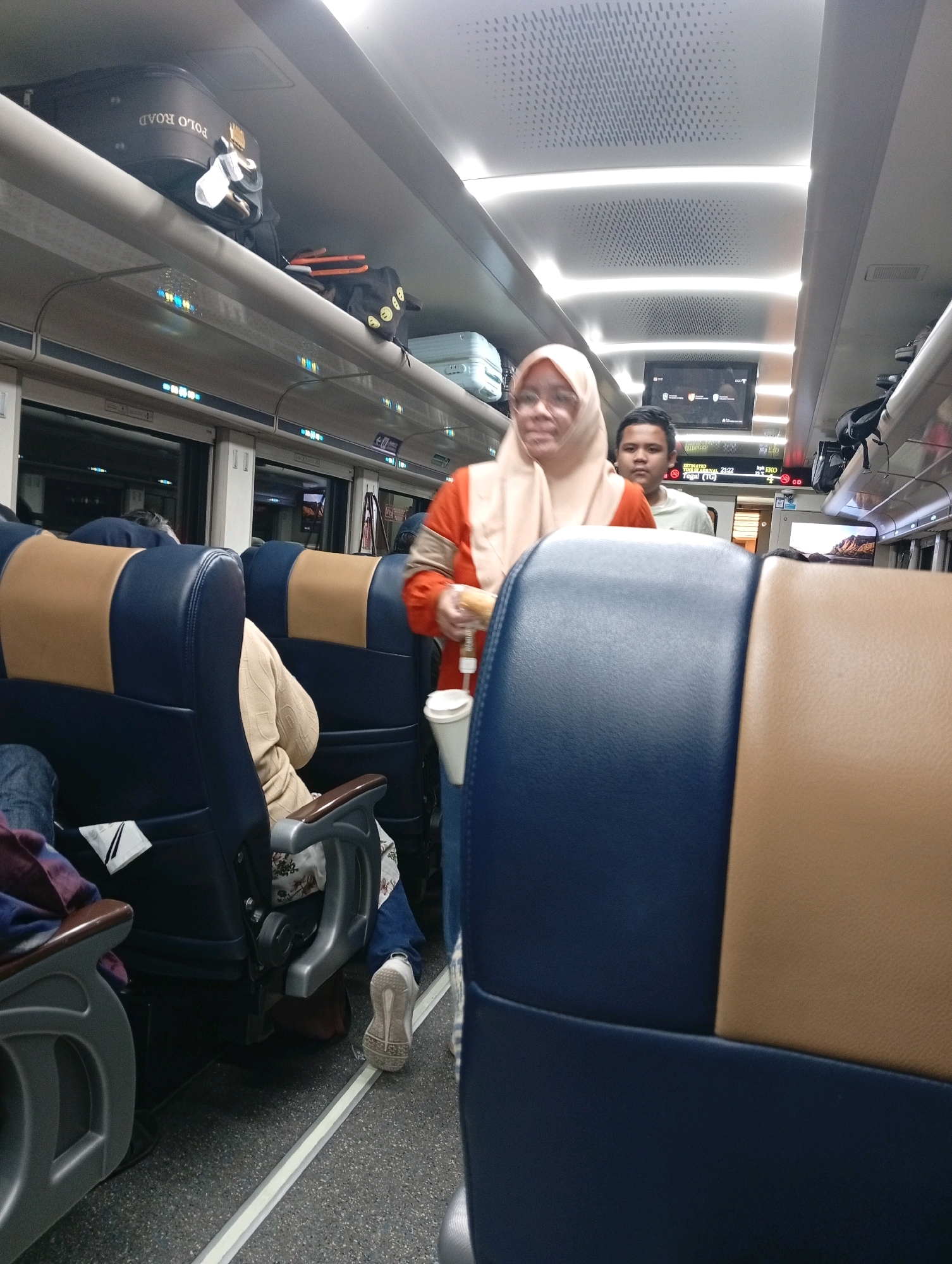 The image depicts the interior of a train carriage, likely in Citemu, Indonesia, during nighttime. Several passengers are visible, including a woman in the foreground wearing a light brown hijab and an orange jacket, holding a white cup. Behind her, a young man in a light green shirt looks towards the camera. The train seats are upholstered in blue and tan. Luggage, including a black "POLO ROAD" suitcase, is stored on the overhead racks. The ambient lighting suggests the train is moving, with illuminated signs and a digital display indicating departure and arrival information. The overall environment is that of a modern train interior.