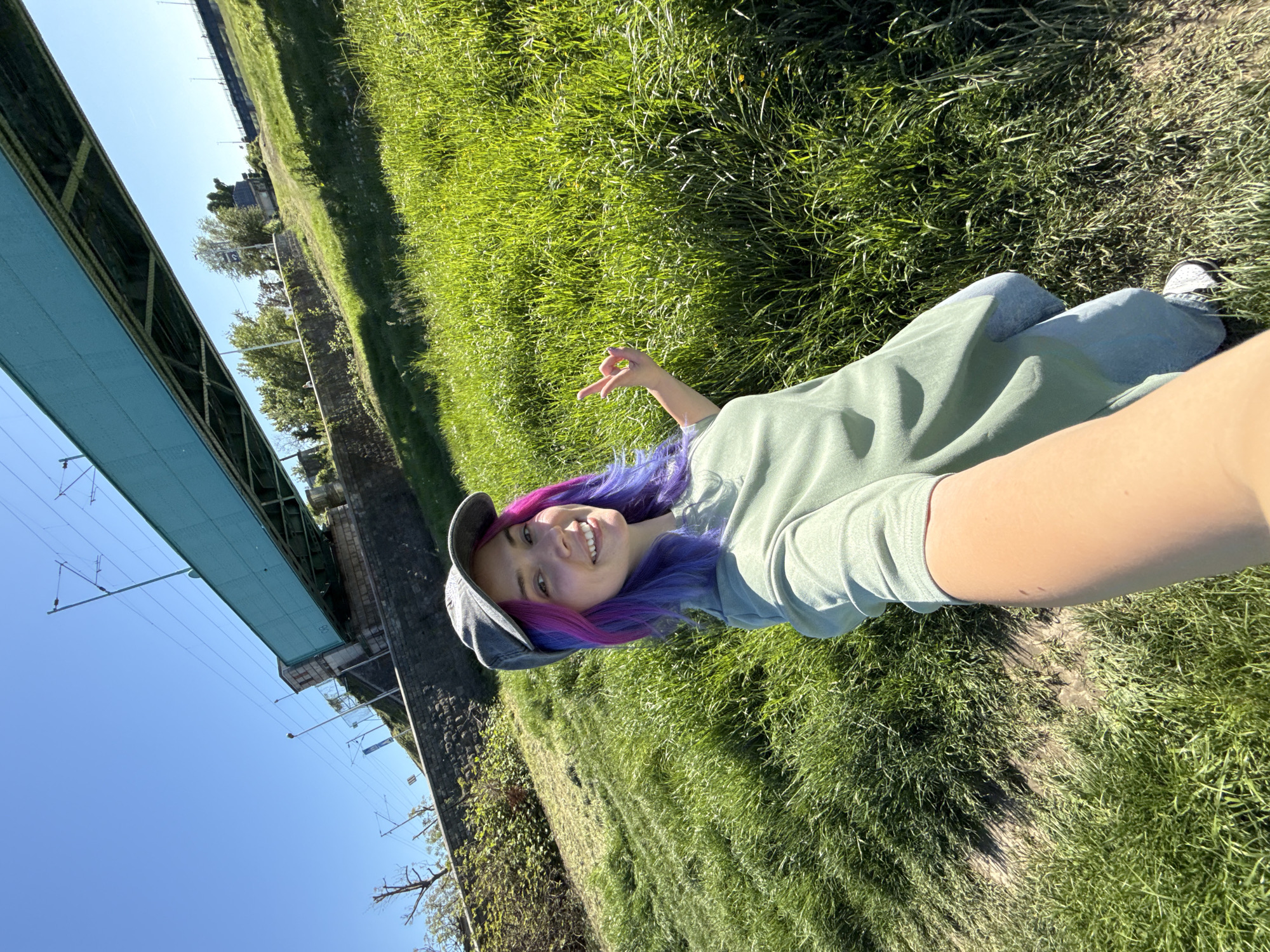 The image is a selfie captured by a young woman lying on her side on a grassy embankment under a clear blue sky. She is smiling broadly and making a peace sign with her left hand. Her hair is dyed in vibrant purple and pink hues, and she is wearing a light green short-sleeved t-shirt, blue jeans, and a gray baseball cap.
The scene is an outdoor setting, likely a park or a natural area adjacent to infrastructure. Most of the foreground and midground is covered in lush green grass, with some patches of bare earth visible beneath and around the person. On the left side of the frame, a prominent light blue metal railway bridge with visible overhead electrical wires spans across the view. On one of the bridge's structures, the number "80" is clearly visible in white. Further in the background, trees and some distant buildings can be discerned.
The activity taking place is a relaxed outdoor selfie. The bright sunshine and clear conditions indicate it is daytime, possibly mid-morning or afternoon. The woman's wide smile conveys a feeling of happiness and contentment. The location is the City of Zagreb, Croatia, implying the bridge is part of the local infrastructure, potentially over a river or a significant railway line in the area.