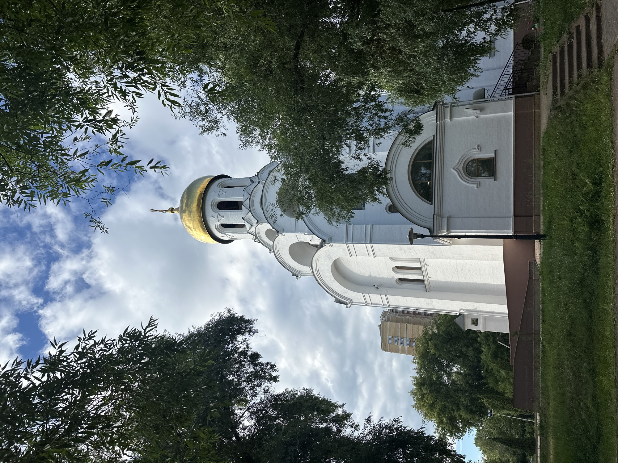 This is a vertical, low-angle, outdoor photograph of a white church with a golden dome. The church has arched windows and decorative architectural details. Lush green trees with dense foliage frame the top and bottom of the image, creating a sense of being under a canopy. Sunlight filters through the leaves, dappling the scene. The sky above is a mix of blue and white clouds, suggesting it is daytime, likely late morning or early afternoon, with partly cloudy weather.
In the background, to the right of the church, a tall, multi-story residential building with numerous windows is visible, indicating an urban or suburban setting. A patch of manicured green grass and a paved pathway run alongside the church. A lamppost stands on the right side of the pathway. The overall impression is one of peace and serenity, with the natural elements of trees and sky complementing the spiritual architecture. There is no visible text in the image. The scene is set in Kotelniki, Russia.