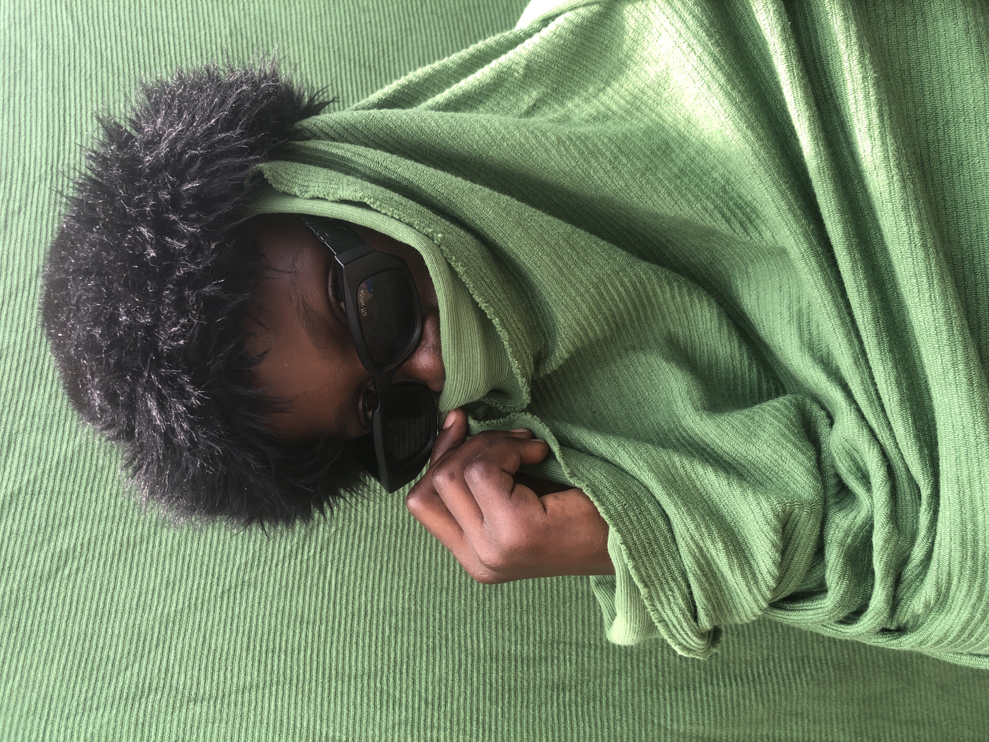 In Ado, Nigeria, a close-up shot focuses on a person's face and upper body. The individual is wearing black sunglasses with "off white" written on them and a fluffy black hat. They are also wrapped in a draped green fabric, which covers most of their face, with one hand peeking out and holding the fabric. The background is a solid green, textured surface, likely a wall or fabric, with horizontal lines. The lighting is direct, casting shadows and highlighting the textures of the fabric and the person's clothing.