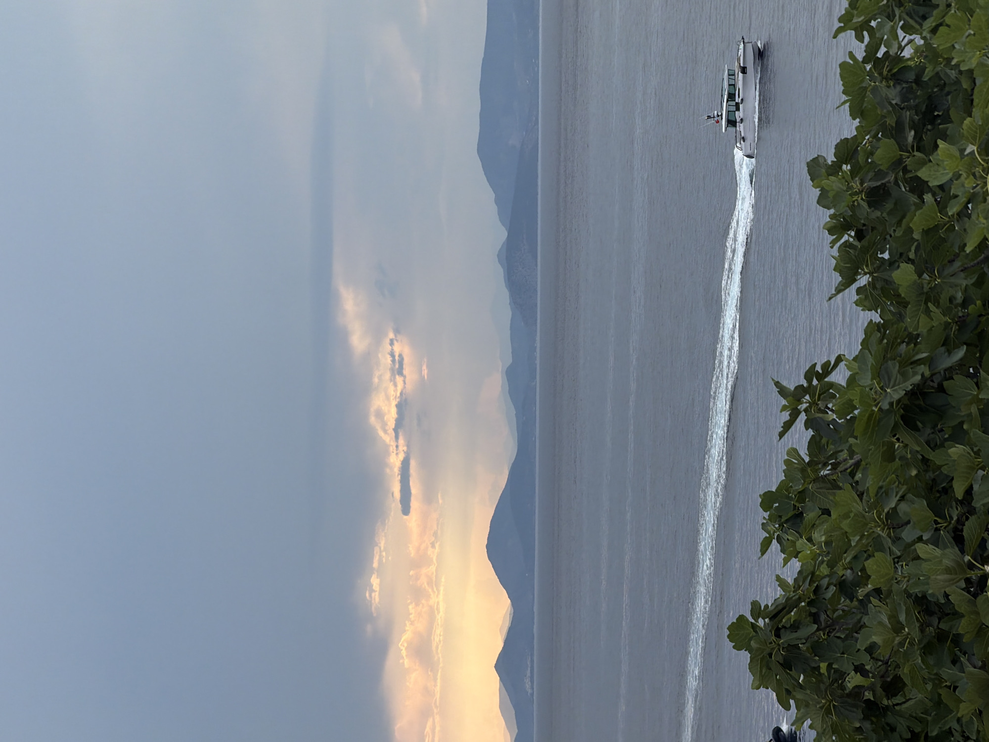 The image captures a boat moving across water near Hydra, Greece. The time is likely late afternoon or early evening, as evidenced by the light hitting the clouds and the shadowed mountains. A boat is visible on the water, leaving a white trail behind it. In the foreground, foliage partially obscures the view of the water and the boat. Mountains line the horizon, and the sky is partly cloudy, with patches of light peeking through.
