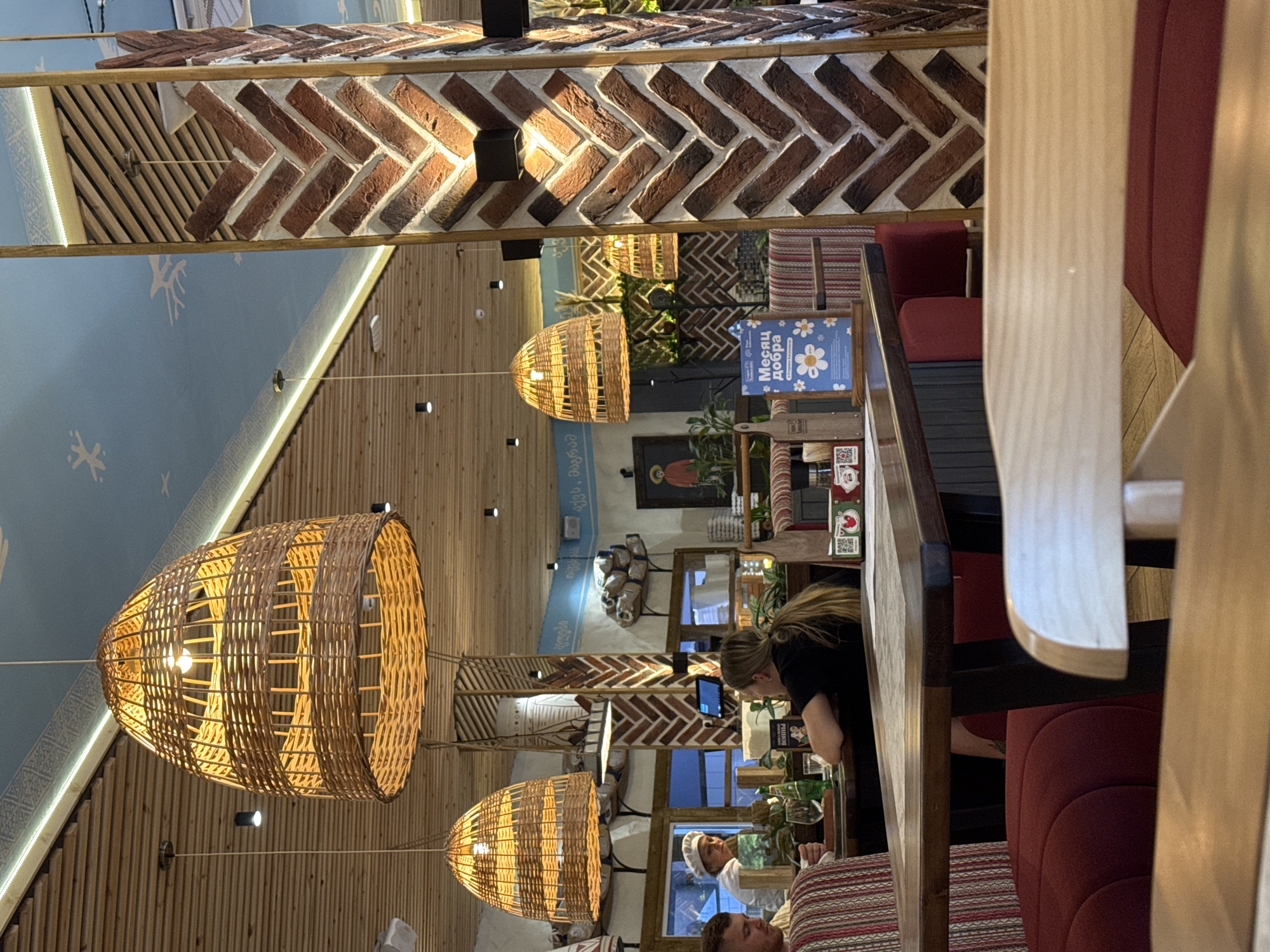 The image depicts the interior of a restaurant, likely in Mikhaylovsk, Russia, given the context provided. The scene is dimly lit and features rustic decor. Dominating the upper portion of the image are large, woven pendant lamps that cast a warm glow. The ceiling is made of wooden planks, and the walls are a combination of exposed brick arranged in a herringbone pattern and painted surfaces with decorative elements.
In the foreground, a dark wooden table is visible, with glimpses of red upholstered seating beneath it. Several people are present. On the left, a man with short hair is seated, wearing a striped shirt. Beside him, a woman with long blonde hair is seated, facing away from the camera, wearing a dark top. In the background, a person wearing a white chef's hat and uniform can be seen behind a counter.
Various objects are scattered throughout the restaurant, including bottles, glasses, and what appear to be decorative items and signage. One sign on a table has text in Cyrillic, and a blue sign with white floral graphics is also visible. The overall atmosphere suggests a casual dining establishment with a focus on a natural or traditional aesthetic.