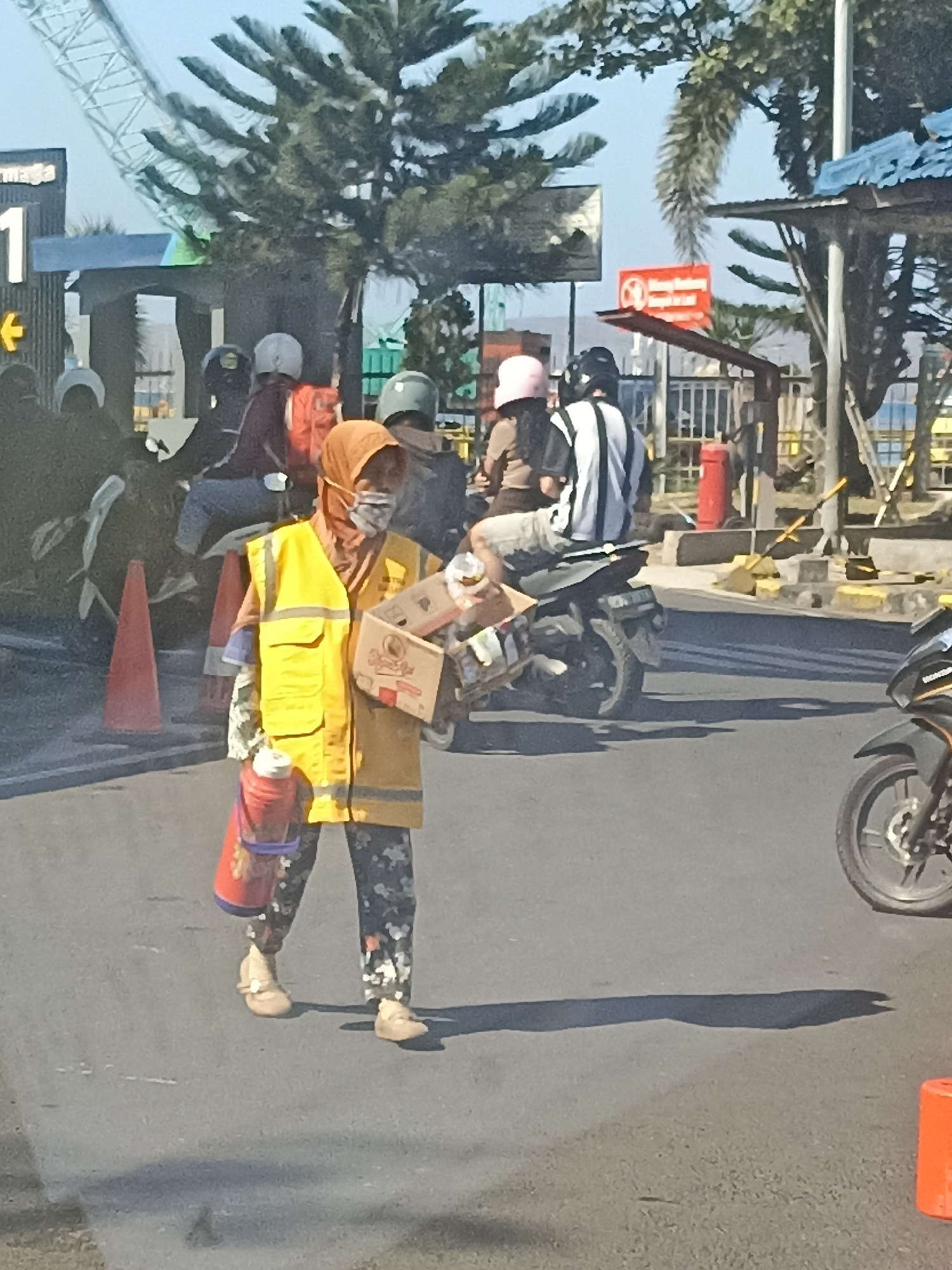 A person wearing a yellow vest, a light-colored hijab, and a patterned face mask is walking on a paved road. The person is also carrying a cardboard box filled with items and a red thermos. The person is wearing light-colored shoes and patterned pants. In the background, there are several motorcycles, some with riders. There are also traffic cones and a large industrial crane visible. The scene appears to be in an urban environment, possibly a street or intersection, with a clear sky. The overall impression is of a busy, daytime scene in Indonesia.