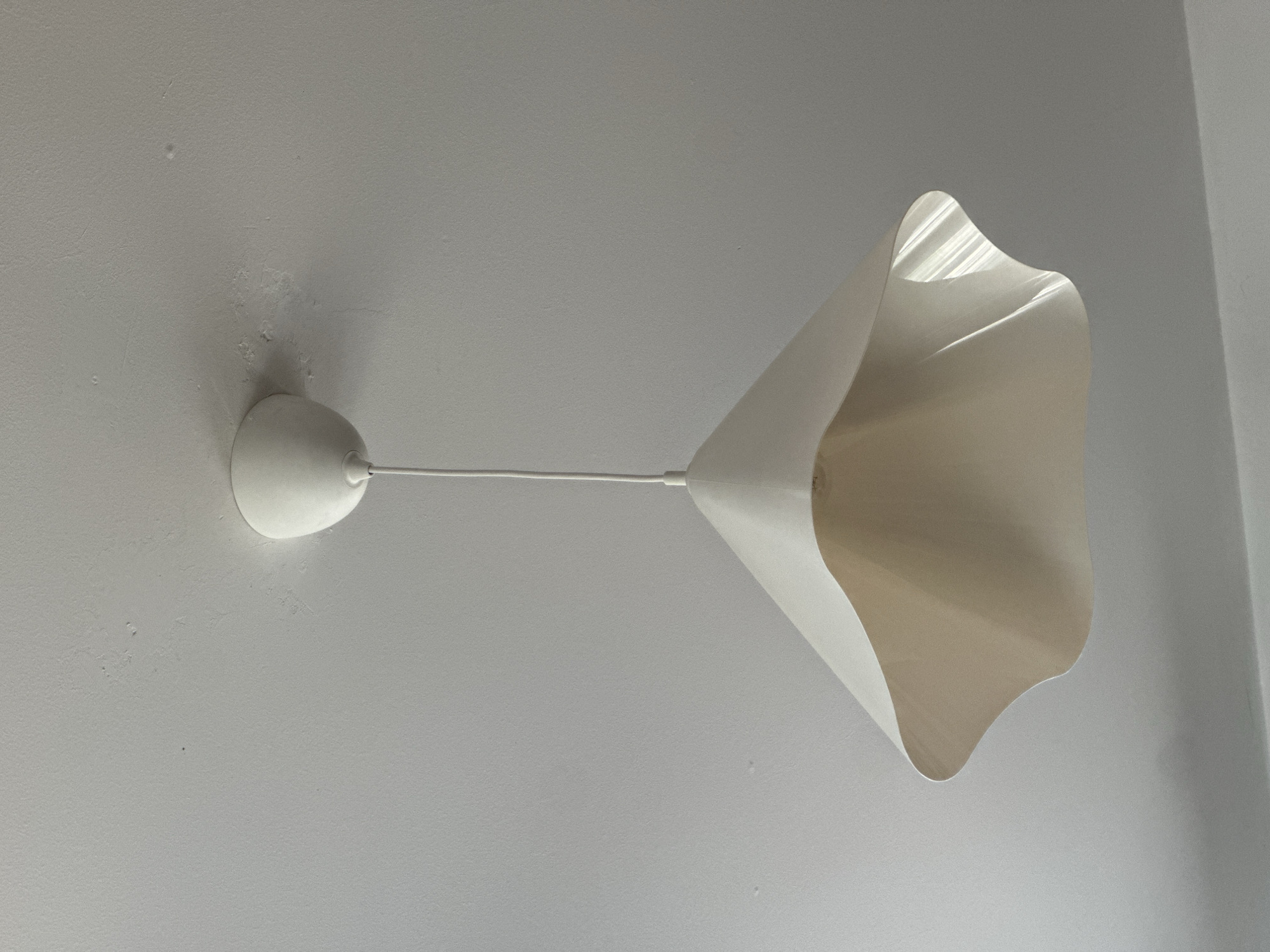 A white pendant lamp with a bulbous base and a long, thin stem terminating in a flower-shaped lampshade is affixed to a white ceiling. The ceiling exhibits minor imperfections, including small raised spots and an area of patching. The image was taken indoors. I cannot locate this specific image in Novi Sad, Serbia.