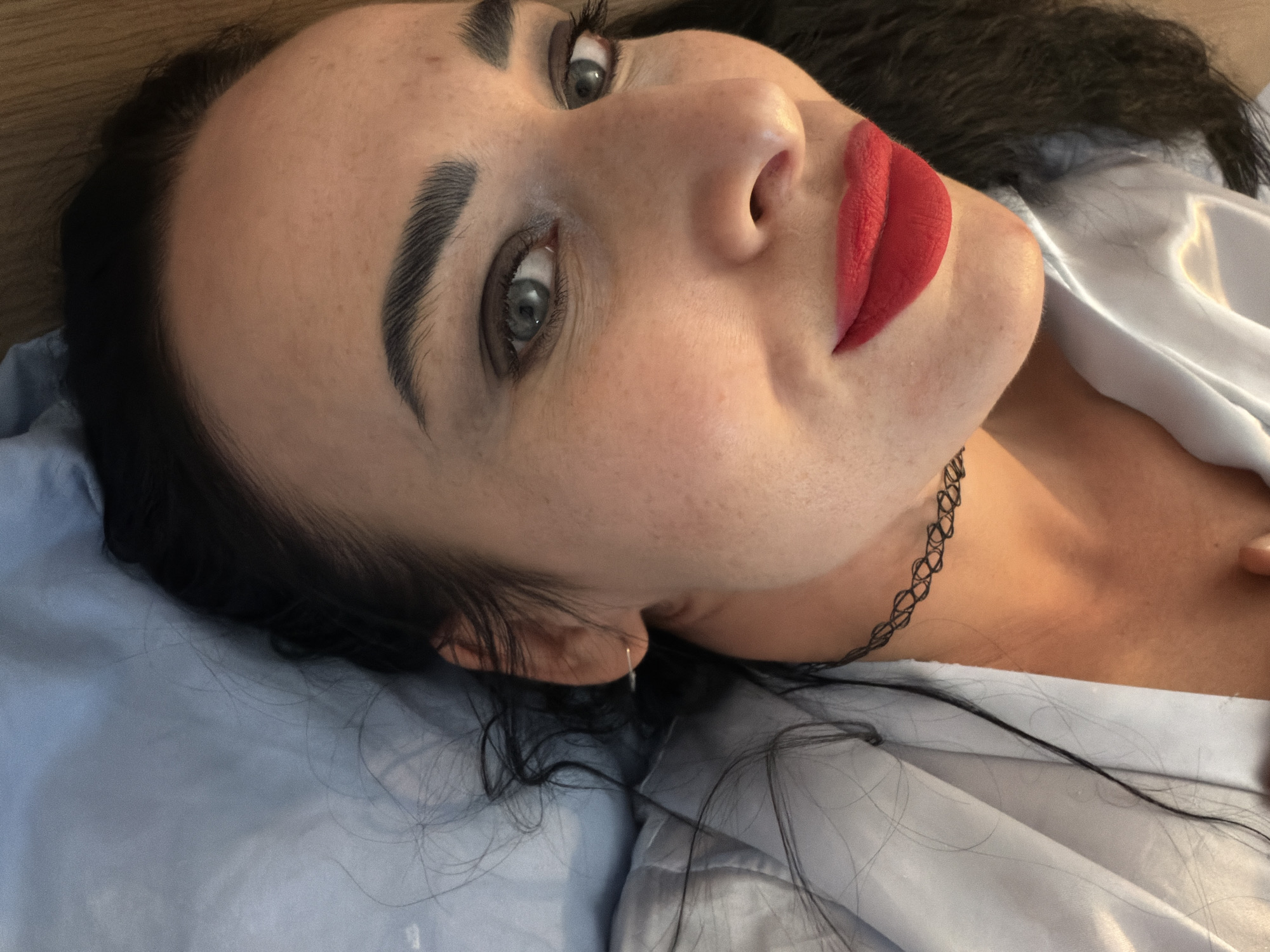A woman with fair skin, dark hair, and red lipstick is lying down. She has dark, defined eyebrows and light-colored eyes. She wears a thin, dark-colored choker necklace and a light-colored shirt. She rests on a blue pillow or bedding. The location is Unknown, Unknown.