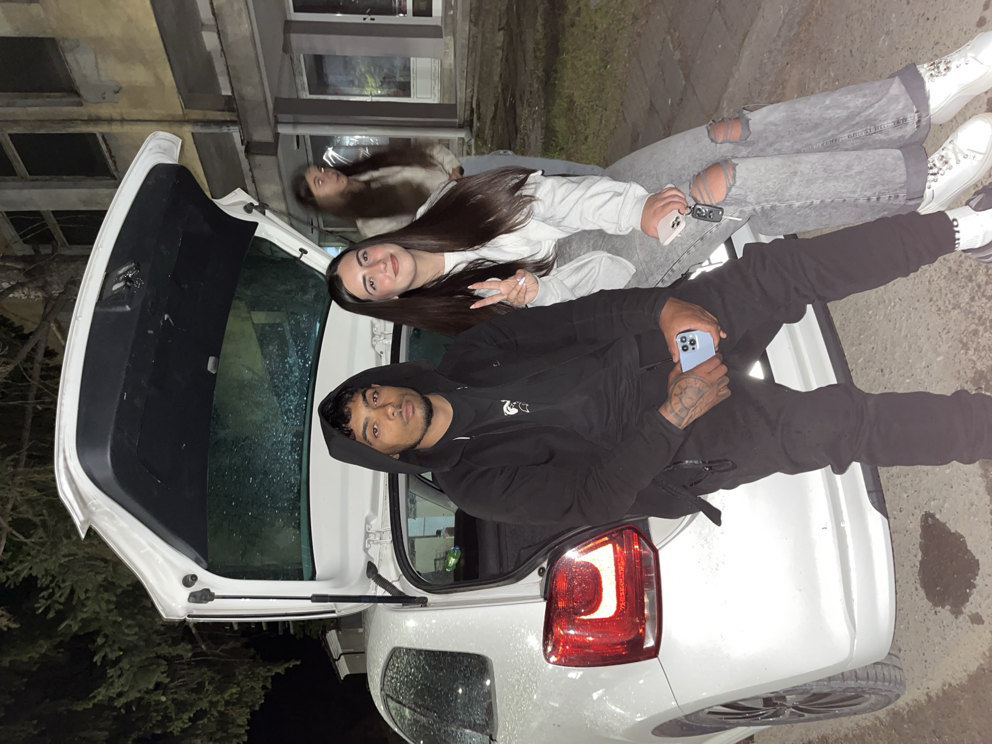 Three people are sitting on the open trunk of a white car. A man in a black hoodie is in the foreground, holding a blue smartphone. A woman in a white hoodie is next to him, making a peace sign with her fingers. Another woman in a grey hoodie and ripped jeans is in the background, looking away. The car is parked on a paved surface next to a building with windows. Vegetation is visible to the left of the car. The location is outdoors at night. The orientation of the image has the scene on its side, making it difficult to discern details. The city of Lozarevo, Bulgaria is provided as background information but isn't visually verifiable.