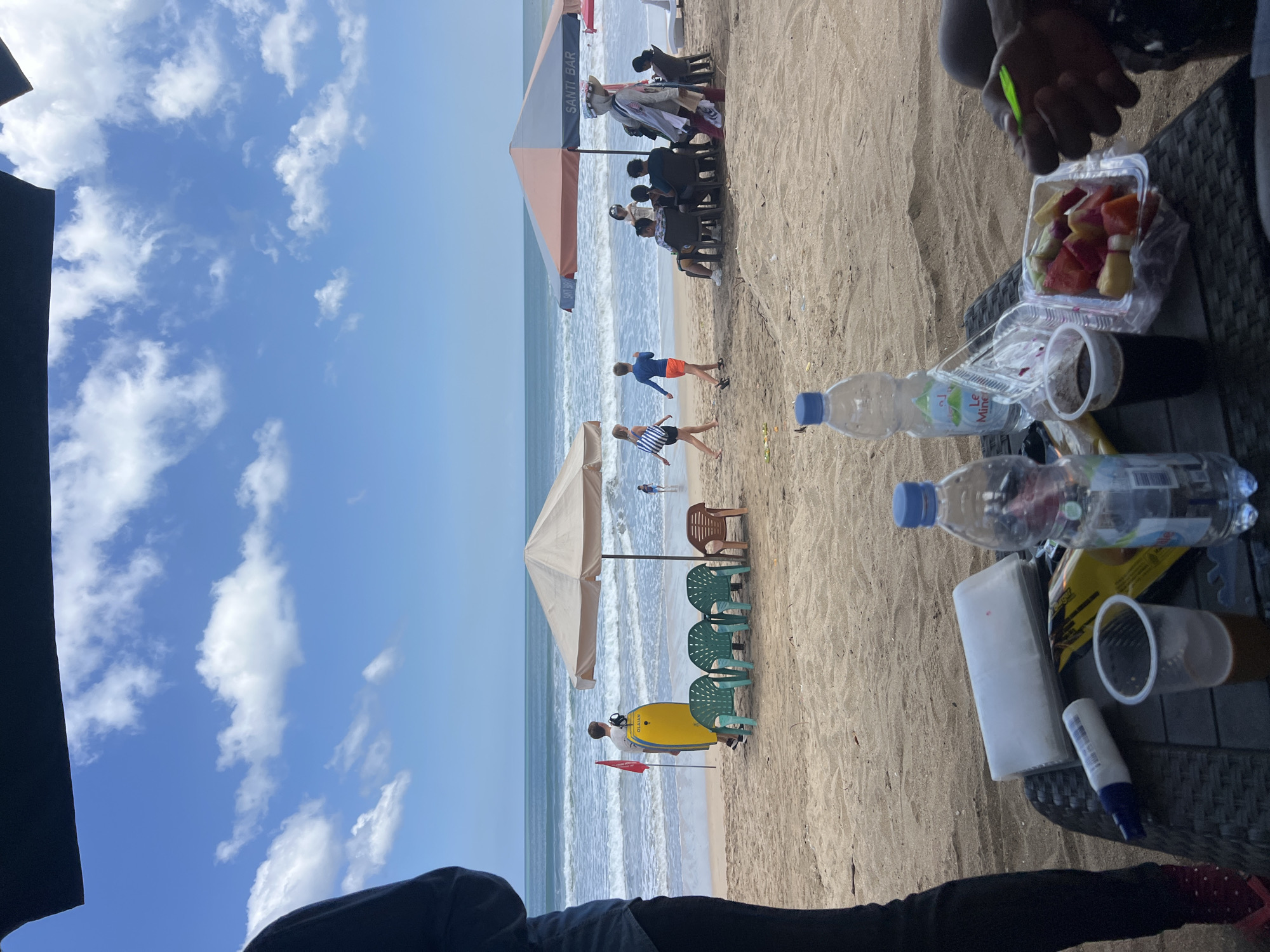 The image depicts a beach scene in Kuta, Indonesia, under a partly cloudy blue sky. In the foreground, a table is set with drinks, a container of sliced fruit, and other items. The table is positioned under an overhang, likely from a beach umbrella or shelter. Beyond the table, the sandy beach is visible with several beach umbrellas, chairs, and people.
To the left, a person stands partially obscured, wearing dark clothing and red sandals. Further back, a man is seen holding a yellow bodyboard, with a red flag and green chairs nearby. A light-colored umbrella shades a few plastic chairs.
In the middle ground, several people are gathered under a larger, two-toned umbrella labeled "SANTI BAR". Some are seated at tables, while others stand. Two children are walking on the beach, one with a striped bag over her shoulder.
The ocean is in the background, with moderate waves rolling towards the shore. Other beachgoers can be seen in the distance, and a few more umbrellas are scattered along the coastline. The overall atmosphere suggests a relaxed day at the beach.