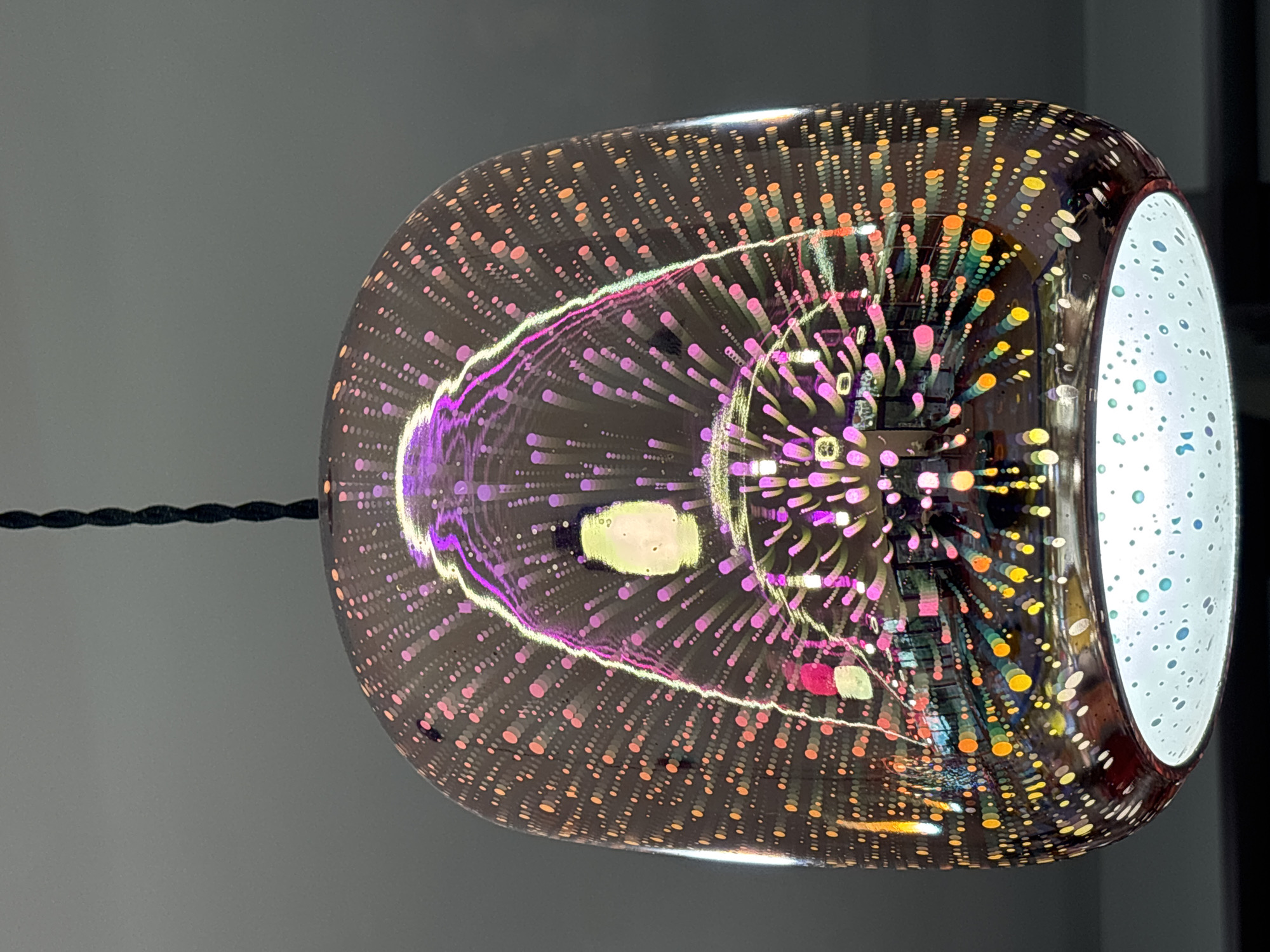 The image depicts a hanging lamp with a decorative glass shade. The shade has a pattern of small colored dots, radiating outward from a central point and giving the illusion of depth. A purple band encircles the central area. The interior of the shade is white with small colored dots. A black twisted cord suspends the lamp. The background is a neutral gray wall. The image was not necessarily taken in Gorna Malina, Bulgaria.