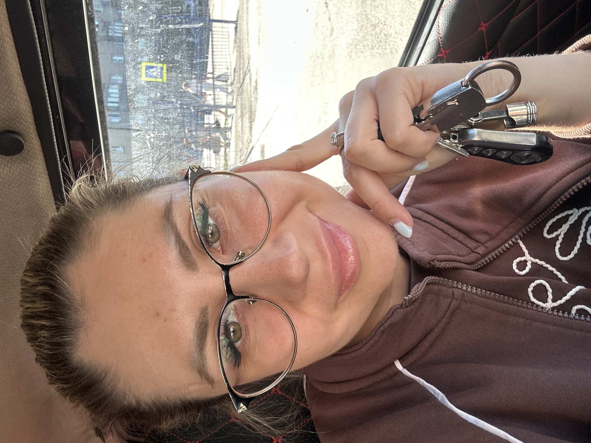 A young woman with blonde hair pulled back and wearing glasses is taking a selfie inside a vehicle. She is wearing a brown zip-up jacket over a brown shirt. Her left hand is resting on her cheek, and her right hand is holding a set of keys. The background appears to be a street scene through the vehicle's windows, with buildings and people visible, suggesting the location is Novoaltaysk, Russia.