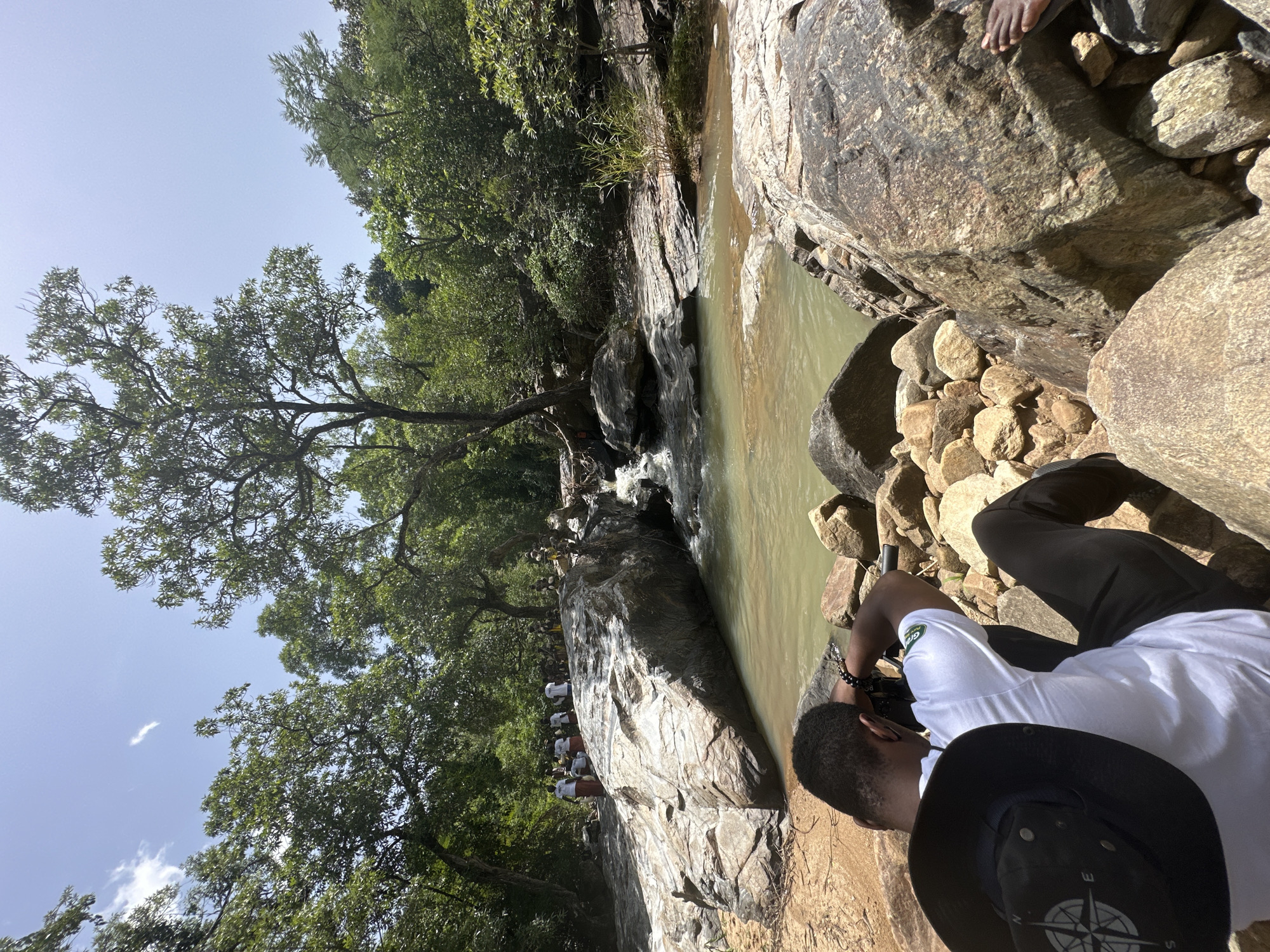 A medium shot captures a person from behind, seated on large rocks by a river in a natural setting, possibly Fobur, Nigeria. The individual is wearing a white t-shirt with a green logo and black pants, accessorized with a black hat featuring a compass design. They are holding a camera, seemingly taking a photograph of the scene.
The environment is lush and green, with dense trees forming a canopy overhead and along the riverbanks. The river itself is a murky, greenish-brown color, flowing over rocks. A cluster of people in white shirts and patterned skirts can be seen in the background, gathered near a large rock formation in the river. The ground is a mix of sandy soil and scattered rocks of various sizes. The sky above is bright blue with a few wispy clouds. The overall impression is of a serene, natural landscape, possibly a destination for tourists or a local gathering.