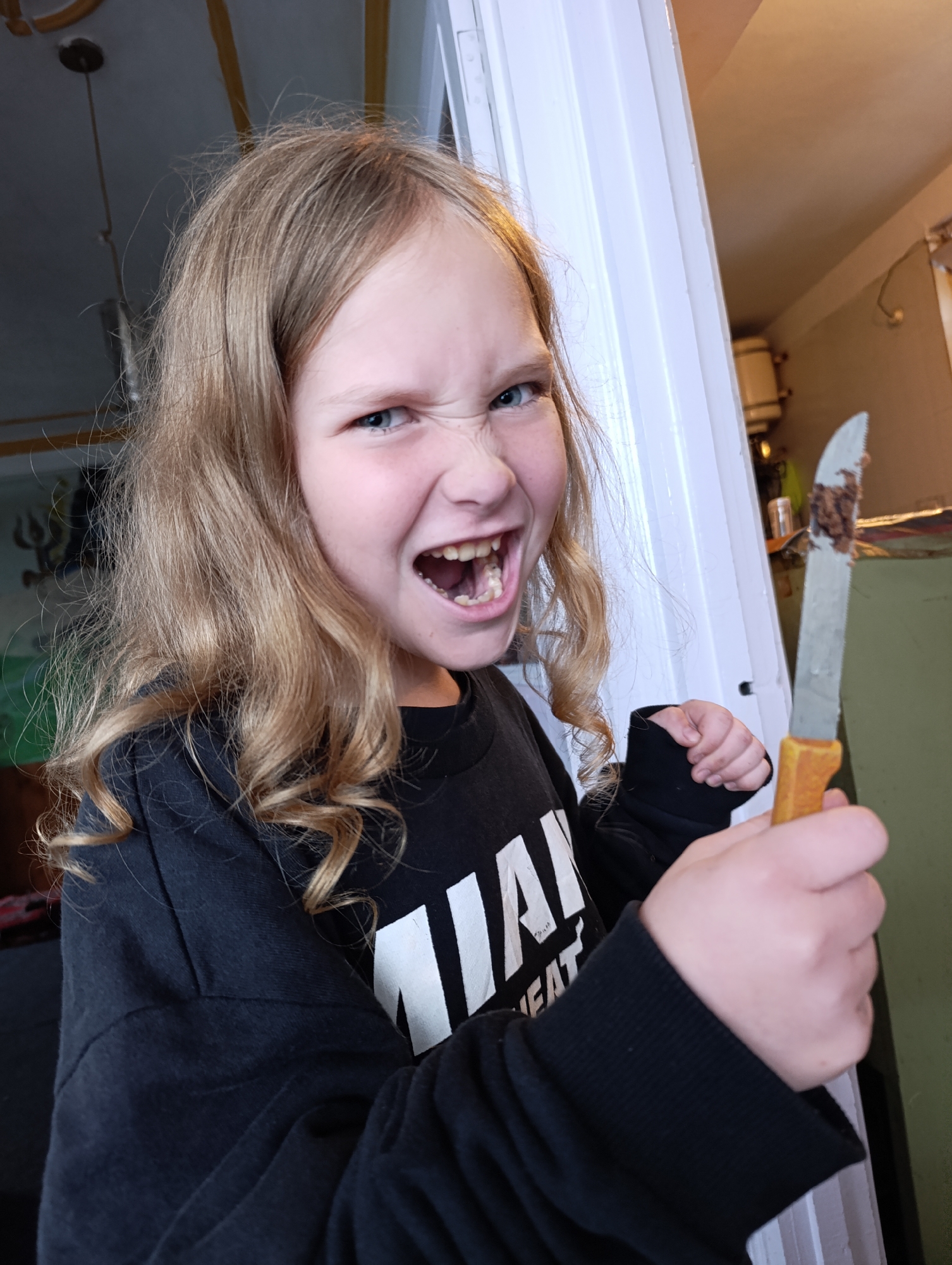 In Vashisht, India, a young girl with blonde, wavy hair and a black sweatshirt with white lettering is captured in a close-up shot. She has an exaggerated, angry expression, with her mouth open and teeth bared. In her right hand, she holds a knife with a yellow handle, its blade smeared with a brown substance. The background is dimly lit and shows a doorway with white trim, and some out-of-focus elements suggesting an indoor environment with some clutter.