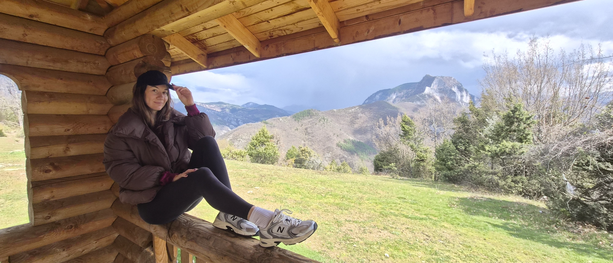 A woman is sitting on the railing of a wooden structure. She is wearing a dark puffer jacket, black leggings, a black baseball cap, and gray athletic shoes with white socks. Her right leg is bent, resting on the railing, while her left leg is straight. She is looking towards the camera and holding the brim of her cap.

The wooden structure appears to be a log cabin or a viewing platform. The background shows a mountainous landscape with green fields and trees. The sky is partly cloudy.

The location is likely Laki, Bulgaria, based on the prompt. The landscape suggests a rural or mountainous area.