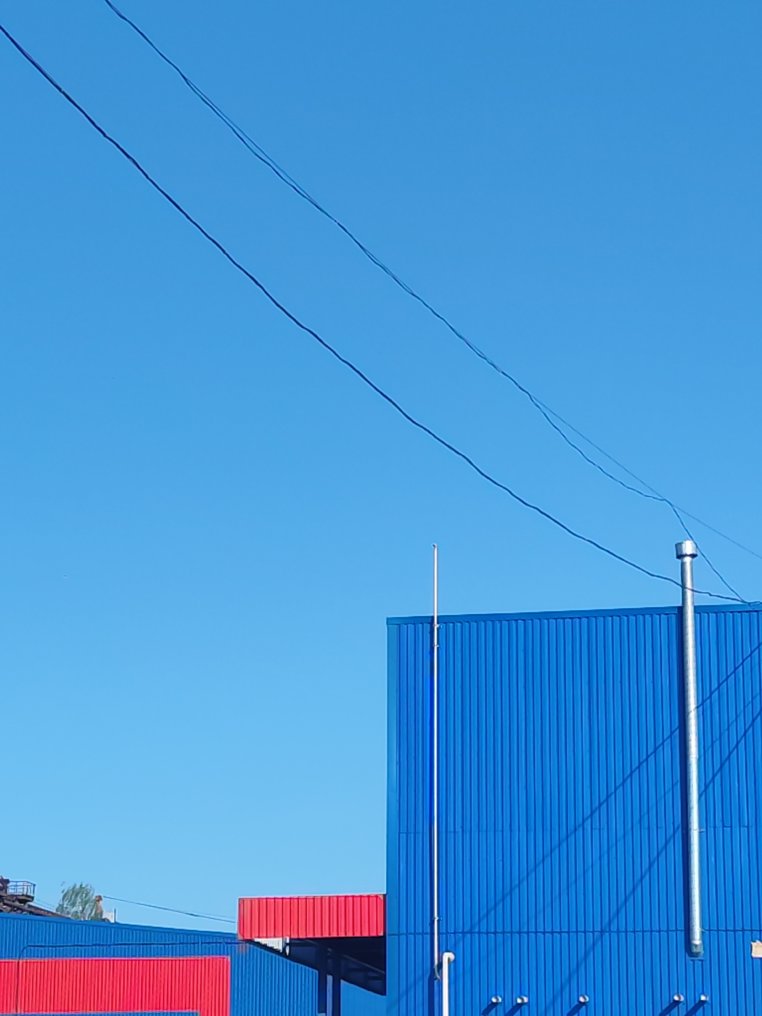 A low-angle shot captures a blue metal building under a bright blue sky with power lines visible in the upper part of the frame. The building features vertical corrugated metal panels, with a silver metal pipe extending upwards along the right side. A thin, rod-like antenna or pole is located closer to the left edge of the blue structure. Elements of a red metal structure are visible near the bottom, extending towards the left side of the image. A portion of a roof with possible foliage peeks in the lower left corner.