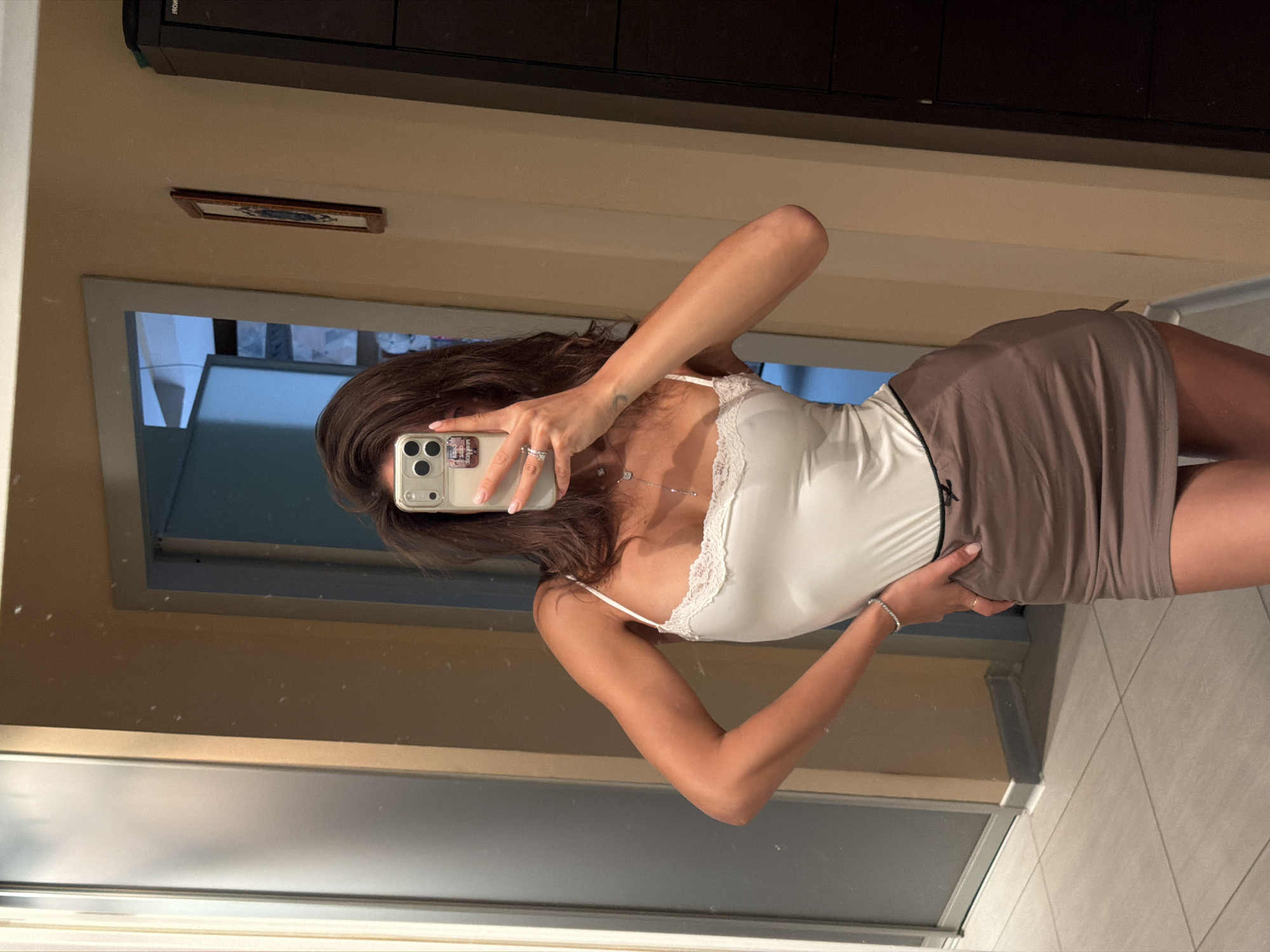 A woman is taking a selfie in a bathroom mirror. She is wearing a white camisole top with lace trim and a taupe-colored mini skirt. She has dark hair and is holding her phone up to obscure her face. Her left hand is visible holding the phone, and her right hand is on her waist. She is wearing rings and a bracelet. The bathroom has light-colored walls, a dark brown cabinet above the mirror, and a framed picture on the wall. The lighting is bright. A window or door is visible in the mirror's reflection, showing a blue and white room. The floor is tiled in light colors. The location in the city of Krasnoyarsk, Russia is not evident in the image.