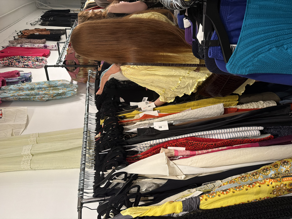 This image depicts an interior retail environment, likely a clothing boutique or second-hand store, situated within Los Angeles, California, United States of America. The scene focuses on two individuals browsing apparel on densely packed racks.
In the foreground, the back of a person with long, straight, auburn hair is prominent. This individual is wearing a loose-fitting, bright yellow top with ruffled sleeves and carries a dark brown, possibly crocodile-embossed, shoulder bag on their right side. Their right hand is extended into a clothing rack, actively engaging with the garments, suggesting browsing or selection.
To the right and slightly behind, the upper back and part of the head of a second person with shorter, lighter brown hair is partially visible, also wearing a dark top.
The central visual elements consist of multiple chrome-plated or silver-colored metal clothing racks densely populated with a diverse collection of garments, primarily on black hangers. The apparel displays a wide range of colors and patterns, including solid black, white, red leopard print, yellow, various shades of blue (striped and solid), cream, and assorted patterned fabrics. Several garments have white price tags attached.
The background features white walls, against which additional clothing items are hung on black wall-mounted racks and directly on the wall. These include a sheer light-green garment, a white shirt with red embroidery, and various patterned and solid-colored tops. The lighting appears artificial and consistent with an indoor retail space.