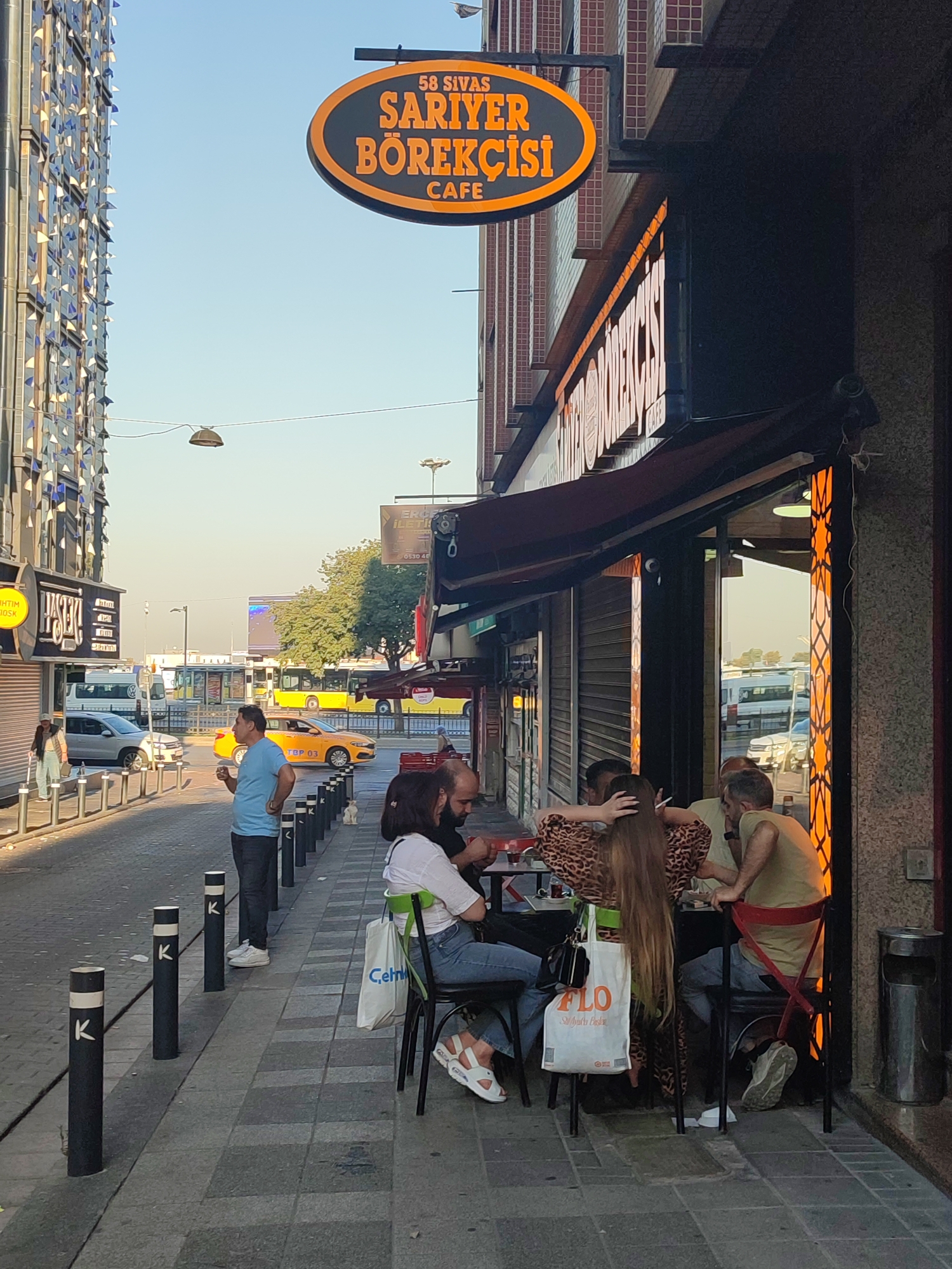 The image shows a street scene in Kadıköy, Turkey, outside a cafe named "58 SİVAS SARİYER BÖREKÇİSİ CAFE." A group of people are seated at tables on the sidewalk, some of whom are interacting with each other. In the background, there is street traffic, including a yellow taxi and a bus, as well as buildings and trees. The lighting suggests it is daytime, likely late afternoon or early evening due to the long shadows. Several bollards line the sidewalk.