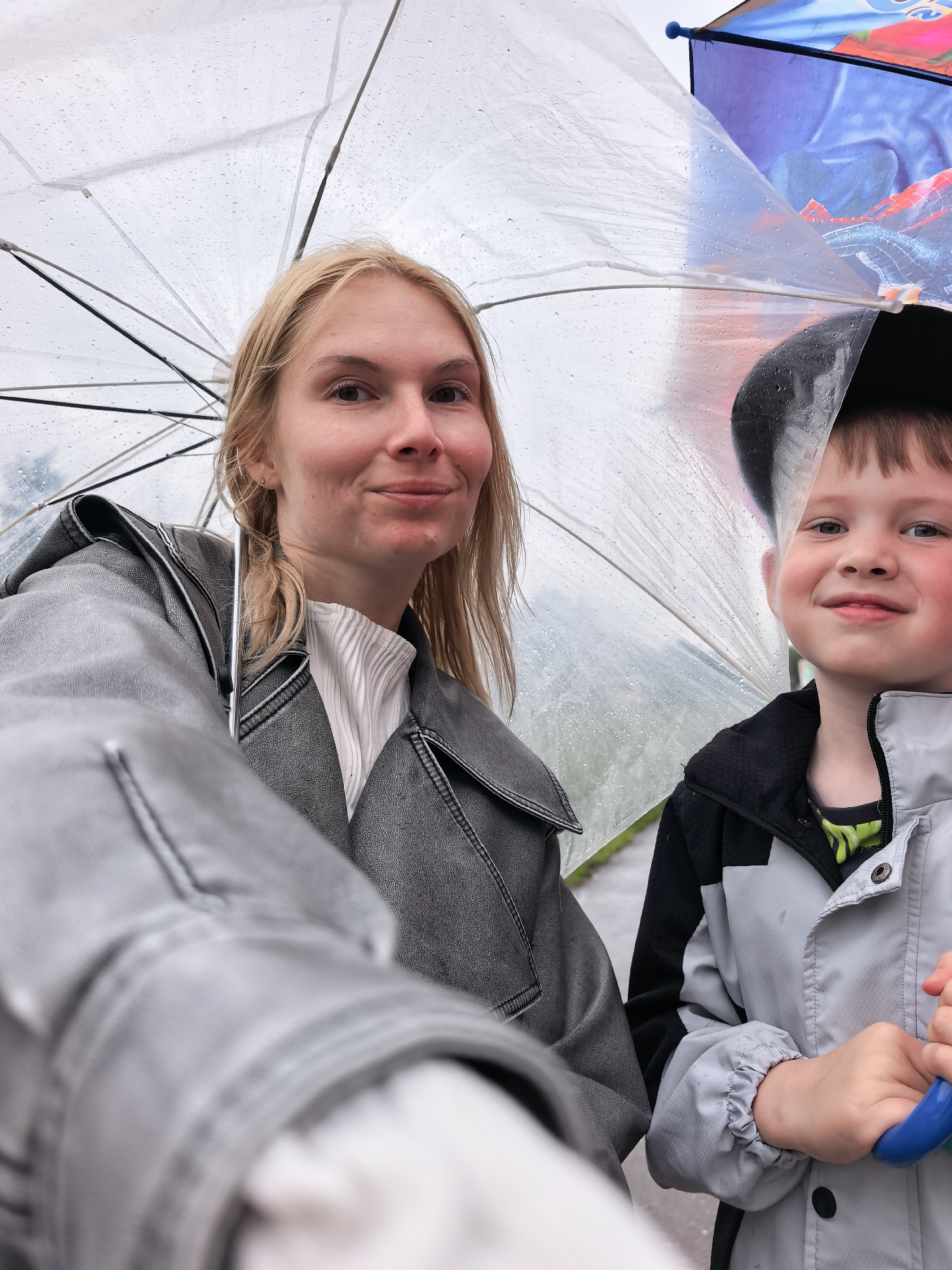 A close-up, selfie-style photograph captures a woman and a young boy outdoors in Sterlitamak, Russia, under umbrellas during rainy weather. The transparent umbrella in the foreground is covered in numerous raindrops, indicating precipitation, and shelters both individuals.
On the left, a woman with blonde hair looks directly at the camera with a subtle smile. She is dressed in a dark grey or black jacket with a leather-like texture, worn over a white, ribbed, high-neck top. Her left arm, extended towards the viewer and partially visible in the bottom left, suggests she is holding the camera.
To her right, a young boy with light brown hair and a black baseball cap also smiles faintly at the camera. He is wearing a grey and black jacket with a zippered front. Behind him, a section of another umbrella is visible, featuring a blue and red design with imagery that strongly suggests the character Spider-Man. A blue tip or finial is also visible at the top of this second umbrella. The boy's right hand, holding a blue handle, is visible in the bottom right, likely the handle of his own umbrella.
The background is softly blurred but indicates an overcast sky and an outdoor setting, consistent with a rainy day in an urban or suburban environment. Both subjects appear calm and content, suggesting a positive mood despite the weather conditions. No discernible text is visible on the subjects or their immediate surroundings, apart from the thematic imagery on the boy's umbrella.