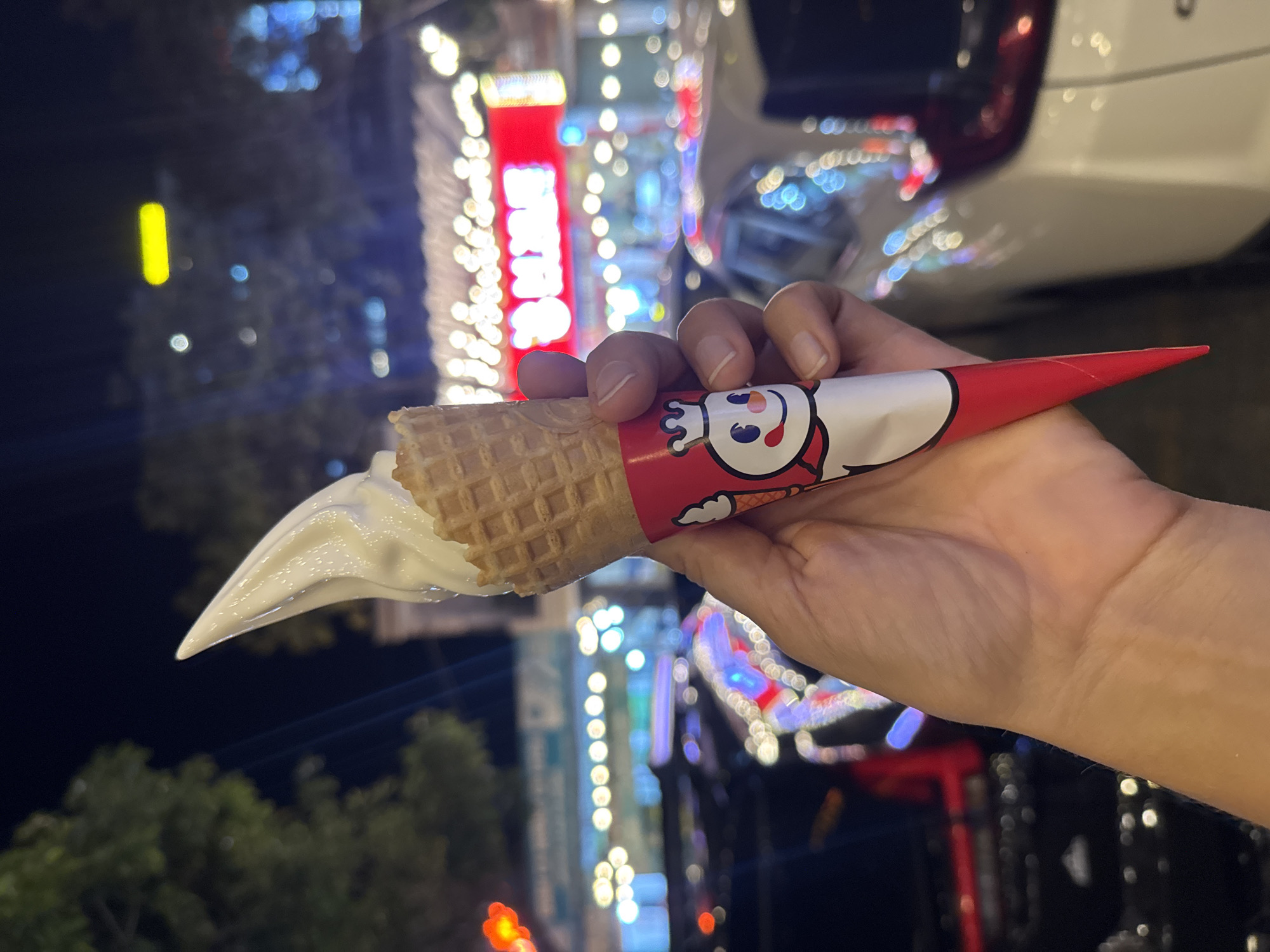 A hand holds a melting soft-serve ice cream cone, wrapped with a red-and-white paper featuring a cartoon character. The background is blurred, showing night-time urban lights and greenery. The location appears to be in the city, and based on the prompt, is in Bac Nha Trang Ward, Vietnam. A car is visible in the upper-right corner of the image.