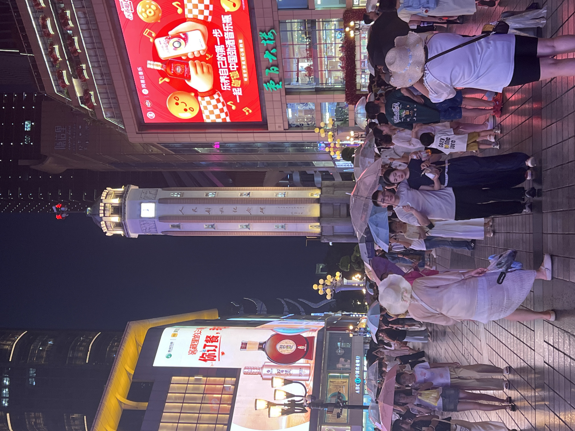 A nighttime scene captures a busy pedestrian street in Yuzhong District, China, likely a commercial area given the presence of large illuminated advertisements and multi-story buildings. A prominent, clock-tower-like structure stands tall, illuminated with lights. The street is paved and wet, suggesting recent rain, with many people walking under umbrellas, indicating active weather. People are seen in various clothing styles, some carrying shopping bags. The advertisements display consumer products, including alcohol and what appears to be a promotional campaign for music. The overall atmosphere is lively and urban, with a mix of contemporary architecture and a significant gathering of people.
