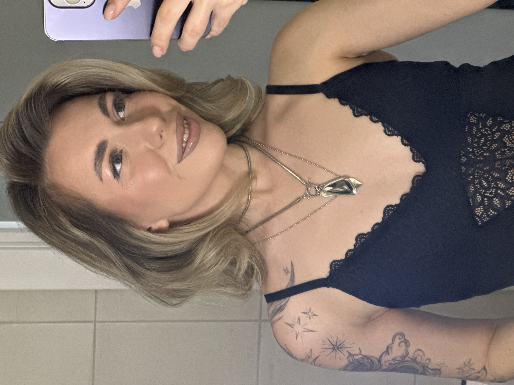 A woman with fair skin, light brown wavy hair, and visible makeup including nude lipstick, is depicted in a mirror selfie. She wears a black lace-trimmed camisole, a choker necklace, and a longer chain with a silver-toned pendant featuring a dark, tear-drop shaped insert. Her left shoulder and upper arm are tattooed with star and celestial designs. She is holding a purple smartphone, possibly an iPhone, in her right hand to capture the image. The background features light-colored tiled walls with dark grout lines and a partial white door frame, indicating an indoor setting. This scene is located in Khimki, Russia.