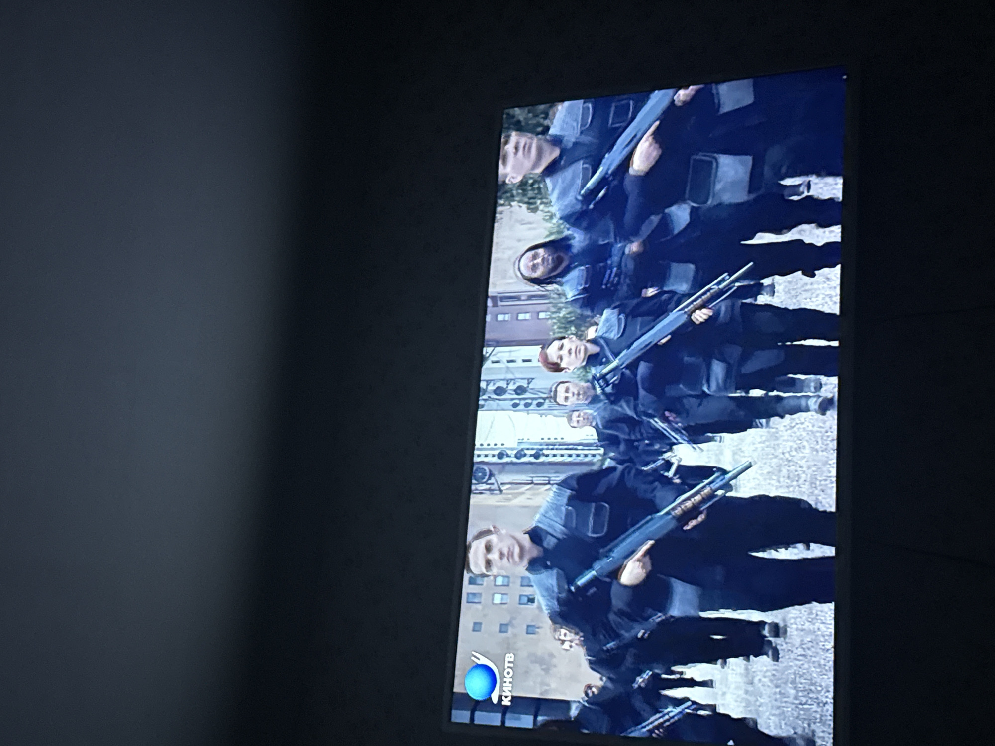 The image shows a television screen displaying a scene from what appears to be a military or law enforcement drama. A group of people in dark uniforms, possibly tactical gear, are holding long firearms. Buildings can be seen in the background, suggesting an urban environment. The bottom left corner of the screen shows a logo, likely for a television channel called “KINOTV”. Based on the logo, the location is likely Mytishchi, Russia.