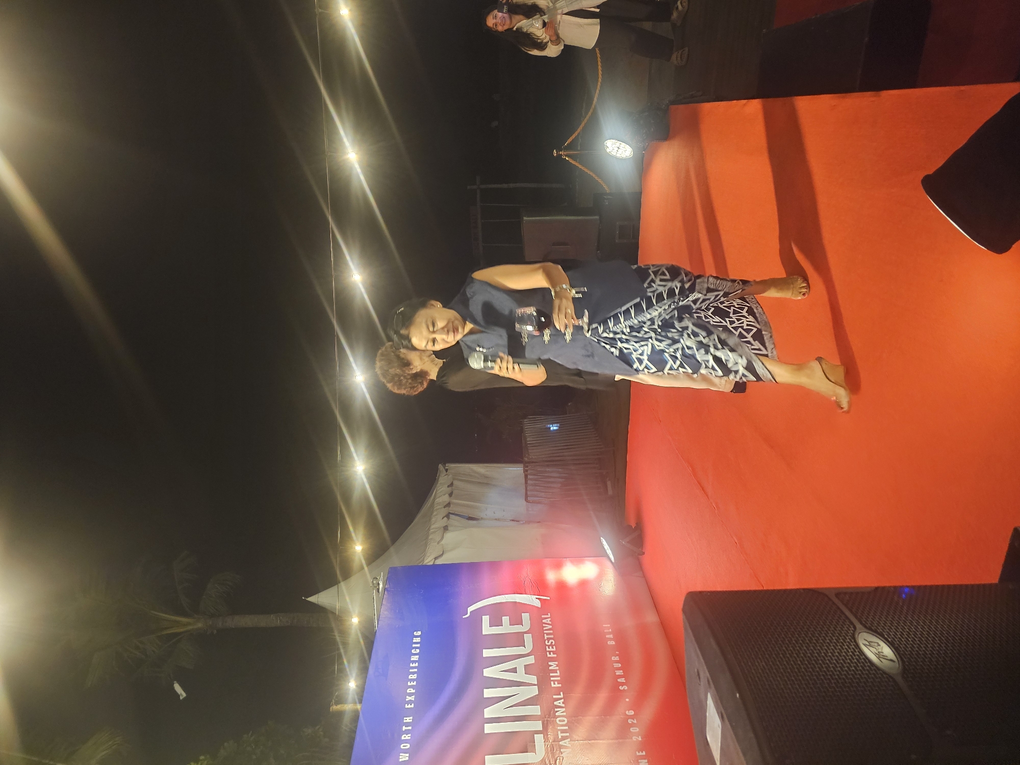 At night in Denpasar, Indonesia, two barefoot women stand on a red platform at an event. The woman on the right, wearing a navy shirt and patterned skirt, holds a microphone to speak. The second woman, to her left and slightly behind, also holds a microphone. A banner is visible behind them with the words "Linale International Film Festival, Sanur, Bali." String lights hang overhead and a palm tree is visible in the background, to the left. A tent is also behind the banner. Some people stand in the distance above the speakers. A black speaker sits in the bottom right corner.