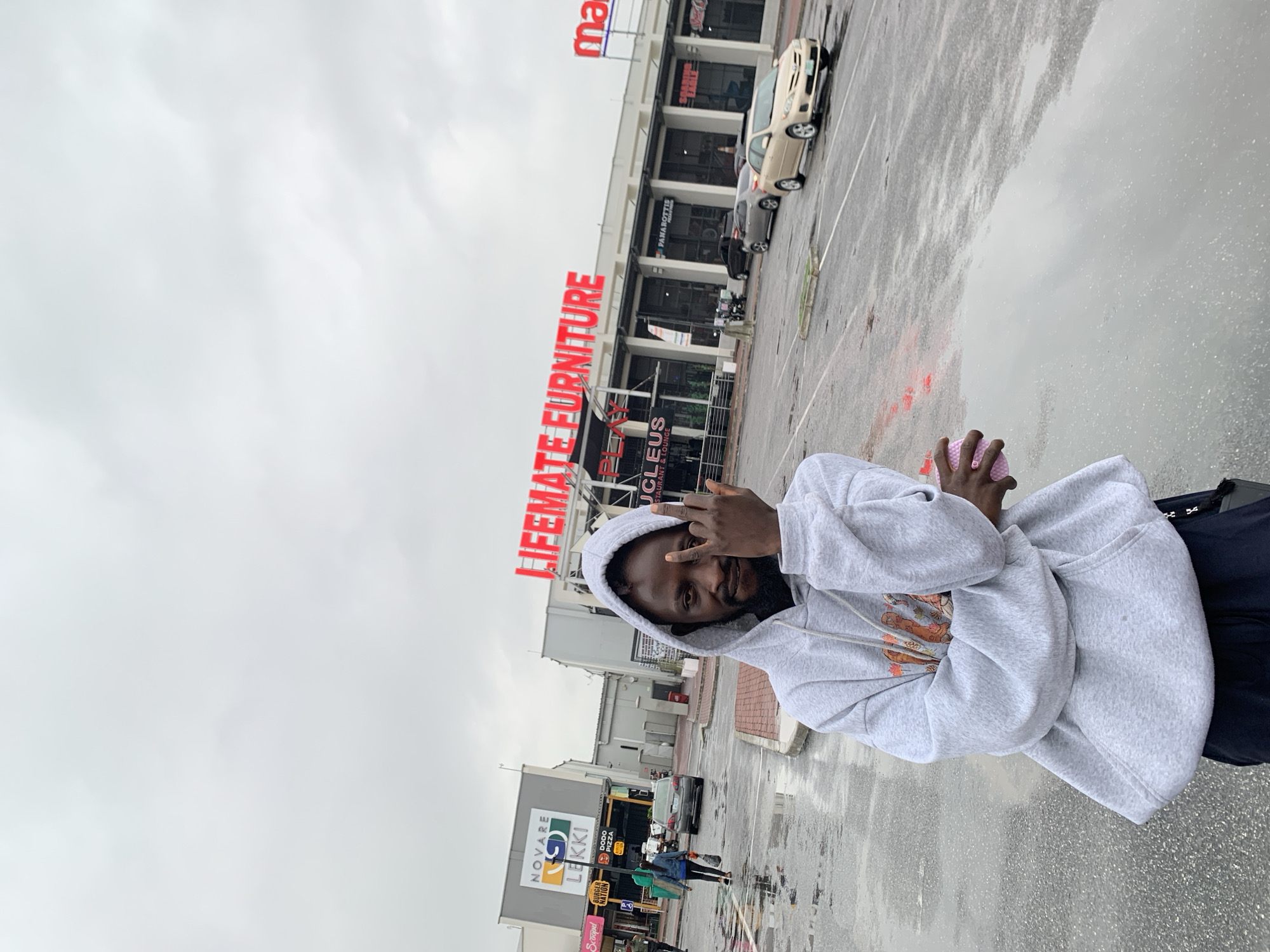 The image captures a person in a gray hoodie standing outside a shopping complex in Sangotedo, Nigeria. The person is facing the camera, holding a pink object in one hand, and making a hand gesture. The shopping complex houses businesses like "LIFEMATE FURNITURE," "UCLEUS RESTAURANT & LOUNGE," "NOVARE," "LEKKI," "BURGER NATION," and "DODO PIZZA." Cars are parked in the complex's parking area, which appears wet from rain. The sky is overcast, suggesting recent or ongoing rain. Other pedestrians are visible in the background near the shopping complex entrance.