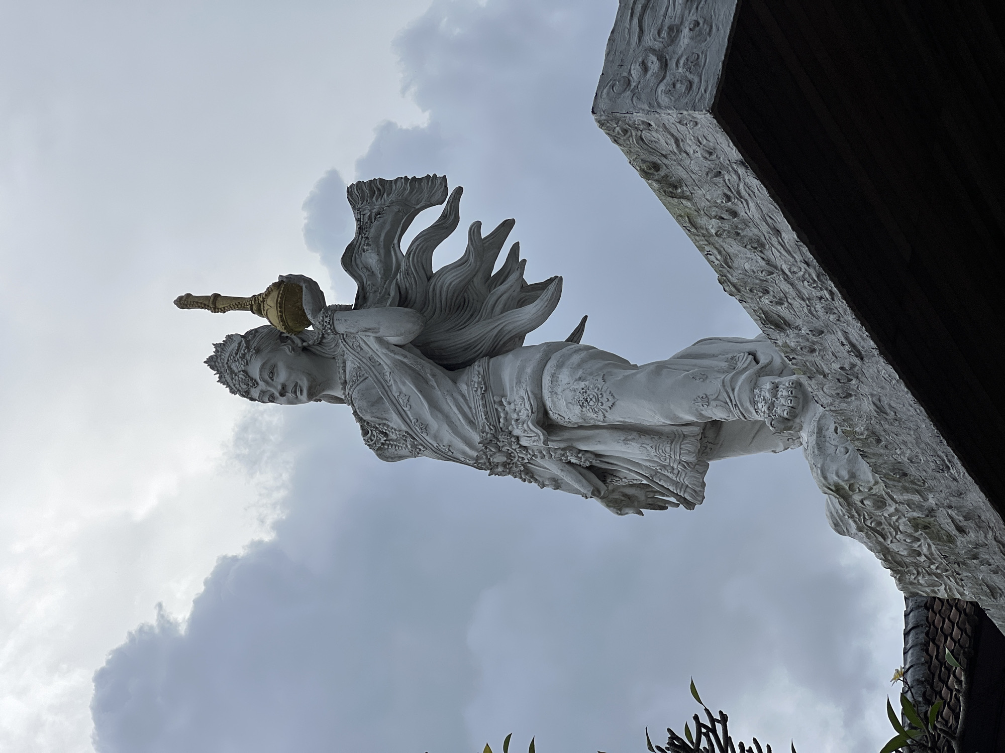 A low-angle shot depicts a white, intricately carved statue of a deity or celestial being, potentially female, against a backdrop of cloudy sky. The statue is positioned as if floating or flying, holding an ornate golden vessel. The deity wears a crown and a flowing garment with decorative elements. A portion of a building structure, possibly part of a temple or other architectural structure, appears in the upper right corner of the image, featuring carved details and dark wooden elements. The location is Celuk, Indonesia, based on the architectural style and cultural context suggested by the sculpture. Vegetation is visible in the lower portion of the frame.