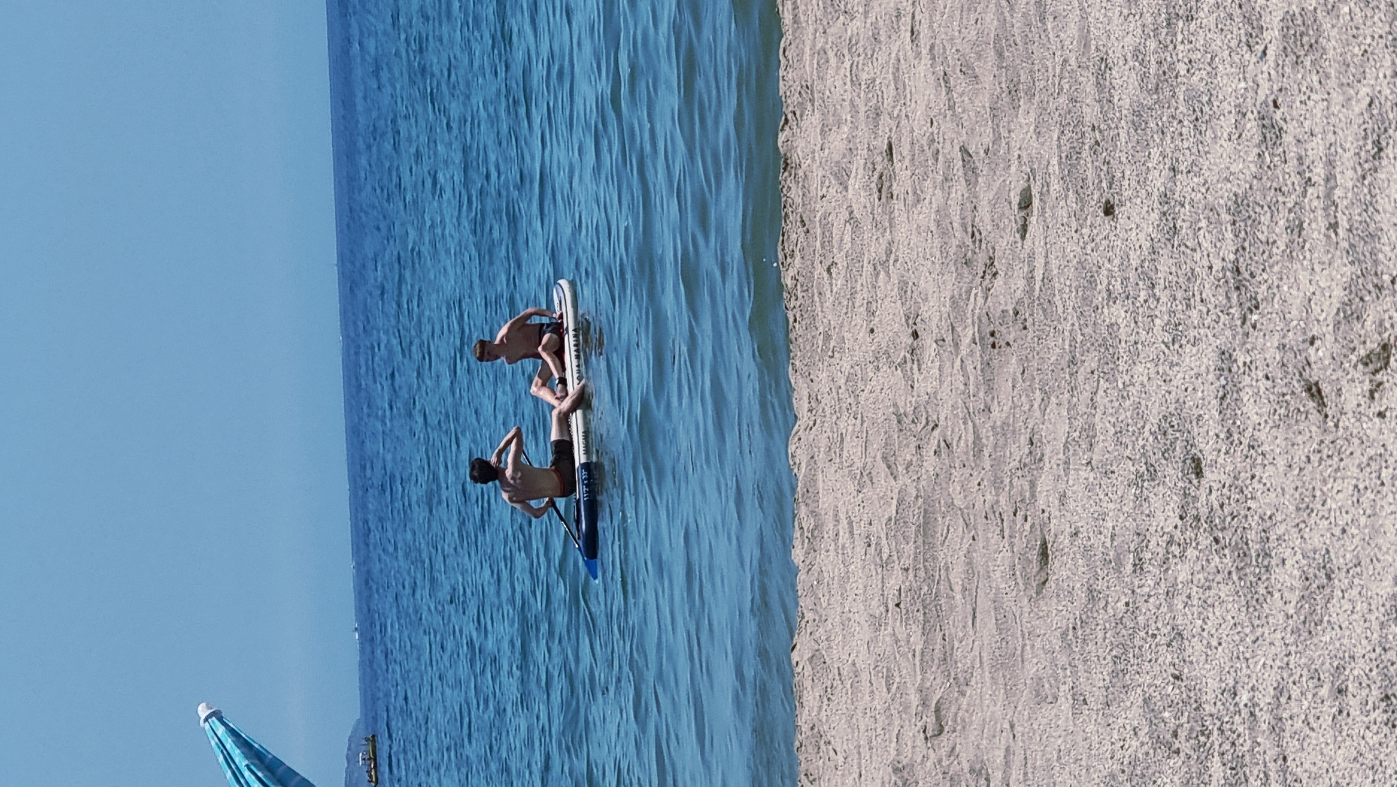 Two young males, appearing to be teenagers, are seated on a white stand-up paddleboard in the calm, blue waters of the Mediterranean Sea. The person on the left is facing away from the camera, looking towards the right, with their right arm raised. The person on the right is also facing away from the camera, looking towards the left. Both are wearing dark swim trunks. The paddleboard has visible markings, including "11'2" x 33"" and "AQUA MARINA." The scene is set on a sunny day, with clear blue skies. In the foreground, a sandy beach with small pebbles is visible. In the distance, a light blue and white striped beach umbrella is partially visible on the far left. The horizon is marked by a distant coastline, possibly featuring some structures. The overall environment suggests a recreational beach setting in Castelló d'Empúries, Spain.