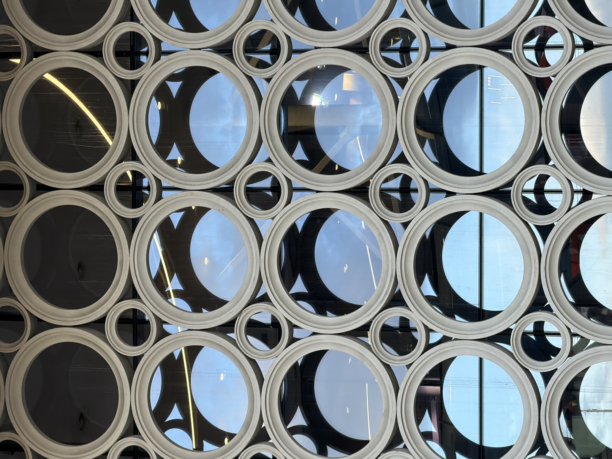 The image shows a close-up, full frame shot of a building facade with a repetitive circular design. The facade is composed of cream-colored, circular frames of varying sizes, arranged in a tessellated pattern. Some of the larger circles are filled with glass, reflecting a blue sky and interior lights. Smaller circular openings are also visible, some appearing as empty frames and others with dark interiors. The overall aesthetic is modern and architectural, with a focus on geometric repetition and light reflection. The location is specified as Unknown, Unknown. There are no people or discernable actions visible in the image.