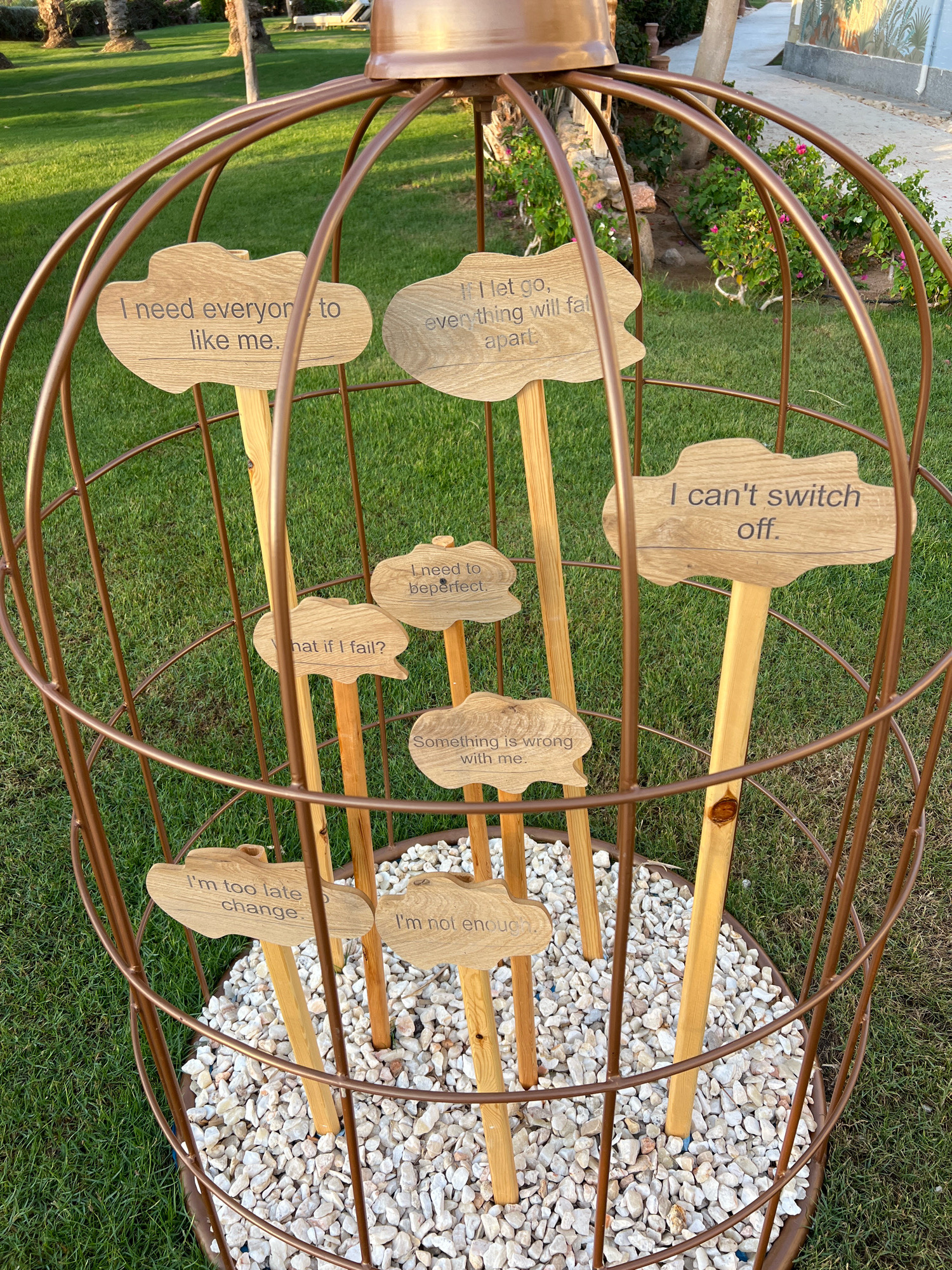 The image shows a large, bronze-colored, birdcage-like structure with a copper-colored dome on top, situated outdoors on a green lawn. Inside the cage, several wooden signs are planted on stakes amidst a bed of white pebbles. The signs are shaped like clouds or speech bubbles and contain engraved text.
The visible text on the signs reads:
- "I need everyone to like me."
- "I let go, everything will fall apart."
- "I can't switch off."
- "I need to be perfect."
- "What if I fail?"
- "Something is wrong with me."
- "I'm too late to change."
- "I'm not enough."
The background shows a well-maintained lawn, palm trees, and some low-lying green plants with pink flowers, suggesting a resort or garden setting. The lighting indicates it is daytime, possibly late afternoon due to the warm tones. The location is identified as Sharm El Sheikh, Egypt. No people are visible in the image.