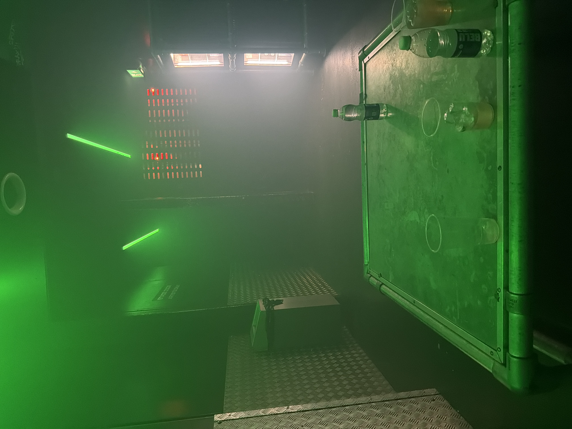 The image depicts the interior of what appears to be a backstage area, possibly in a theatre or performance venue. The overall lighting is green-tinted and hazy, creating a slightly disorienting atmosphere. Visible elements include a metal-grated floor, a box-like structure on the floor, and a panel attached to the wall which has cups and water bottles adhered to its surface. Two green fluorescent tubes are on the wall. Above the horizontal panel are small windows, and a wall panel with small red lights which appear to be ventilation or a lighting fixture. Text that reads "STAGE DOOR ARTISTS ONLY" is partially visible on the wall. No people are visible in the image. While no precise location within London is given, the presence of a backstage area in the context of the United Kingdom suggests a London theatre or performance venue is a likely setting.
