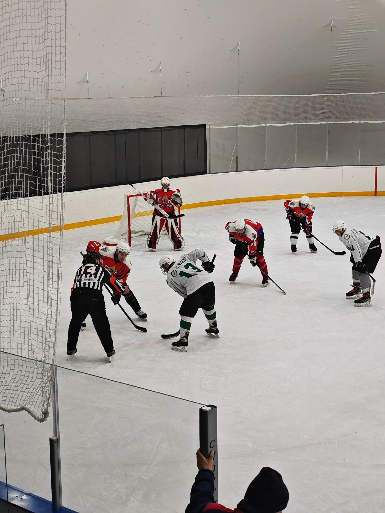 The image depicts a hockey game in progress. Players in red and white uniforms are engaged in a face-off against players in white and green uniforms. A referee in a black and white striped uniform is positioned between them. A goalie in a red and white uniform is standing in front of the net. Spectators are visible in the lower portion of the image. The arena appears to be indoors. The goalie is wearing a uniform that says "COK ПИТЕР" which is likely an abbreviation for "Saint Petersburg Sokol" suggesting that the game is in Saint Petersburg, Russia.