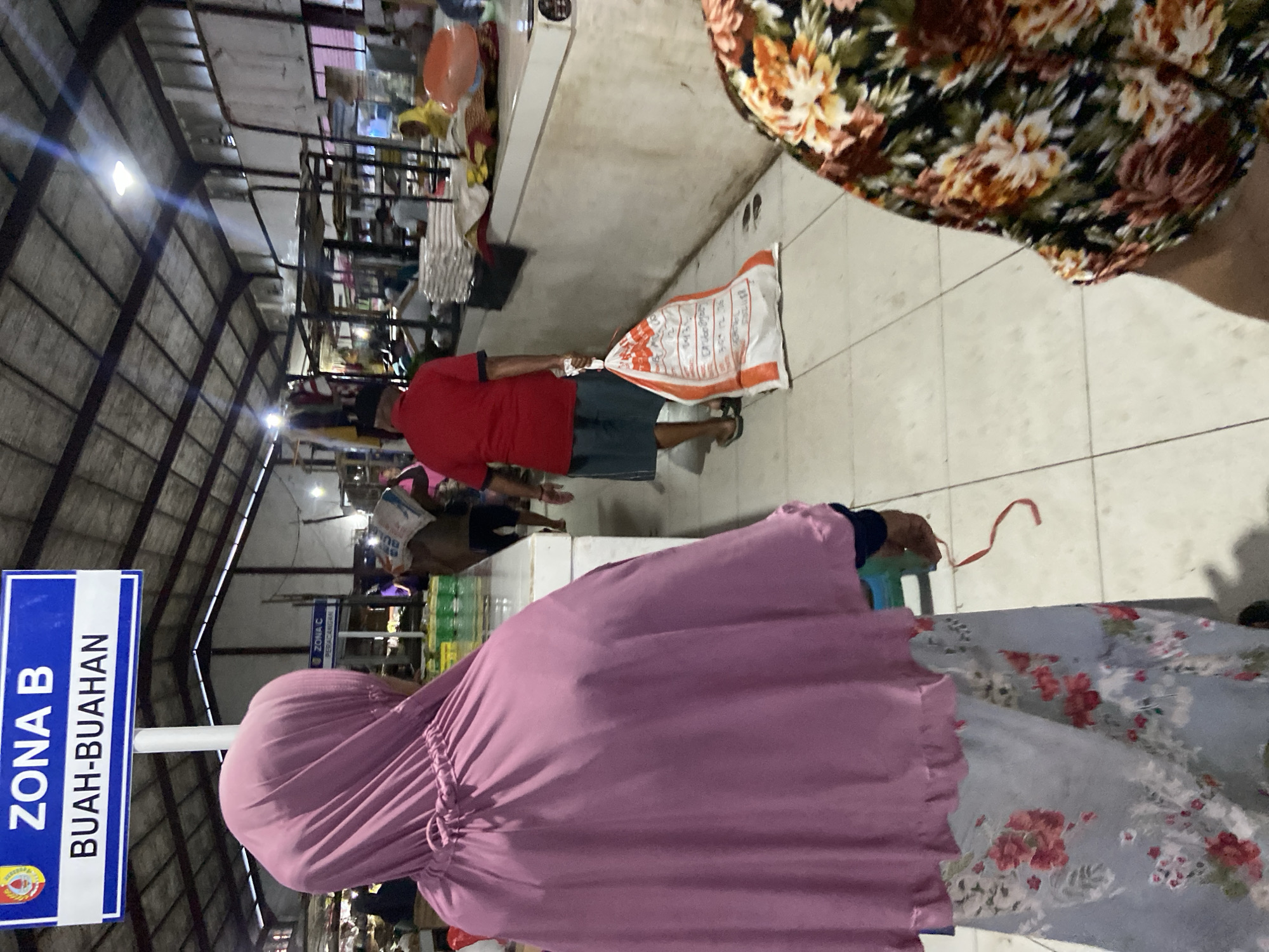 The image shows an indoor market in Jember, Indonesia. The scene is composed of several people with shopping bags, a fruit zone sign, and a warehouse-like roof. A woman with a pink hijab and floral dress is in the foreground and another woman in a floral top is on the right, partially obscuring the view. A man wearing a red shirt, dark shorts, and a hat walks with a white sack in the center of the scene. Other figures are in the background, engaged in shopping or market activity. The sign on the left side reads "ZONA B BUAH-BUAHAN", indicating the location of the fruit section. The area is lit by overhead lights, typical of indoor market settings.