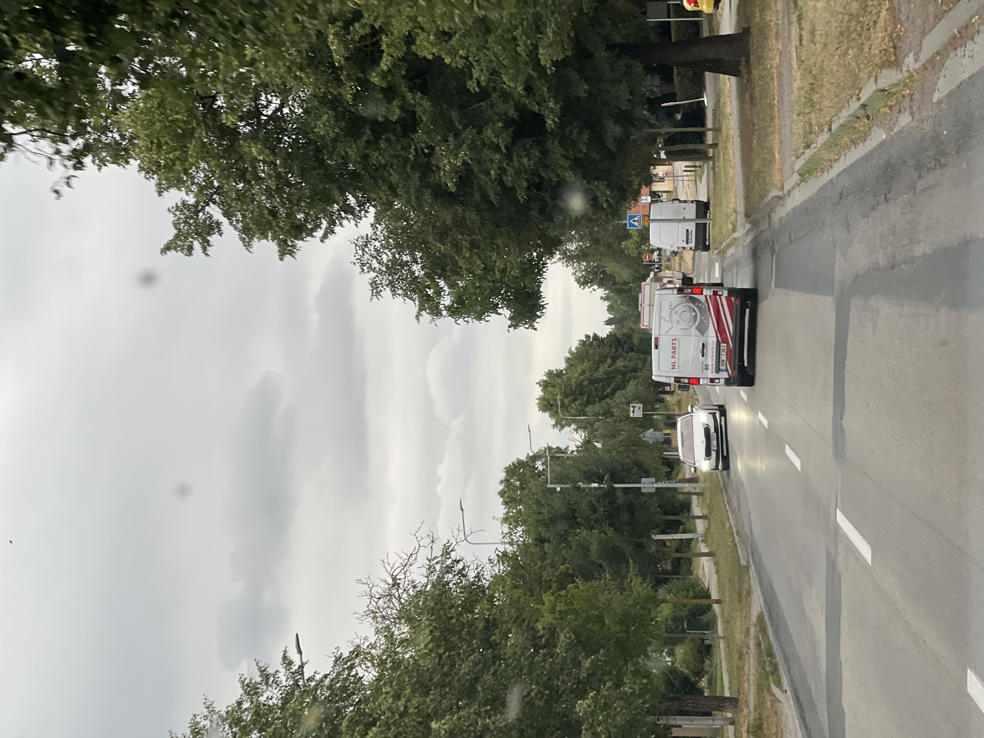 The image shows a street scene in Měnín, Czechia. The weather is overcast. On the right side of the road, a row of green trees lines the sidewalk. A white van with red and white decals that reads "ML PARTS" is driving in the middle lane, followed by another white van. A white sedan is in the left lane. Further down the road, there are more trees and buildings. The road has white lane markings. There are streetlights visible along the road.