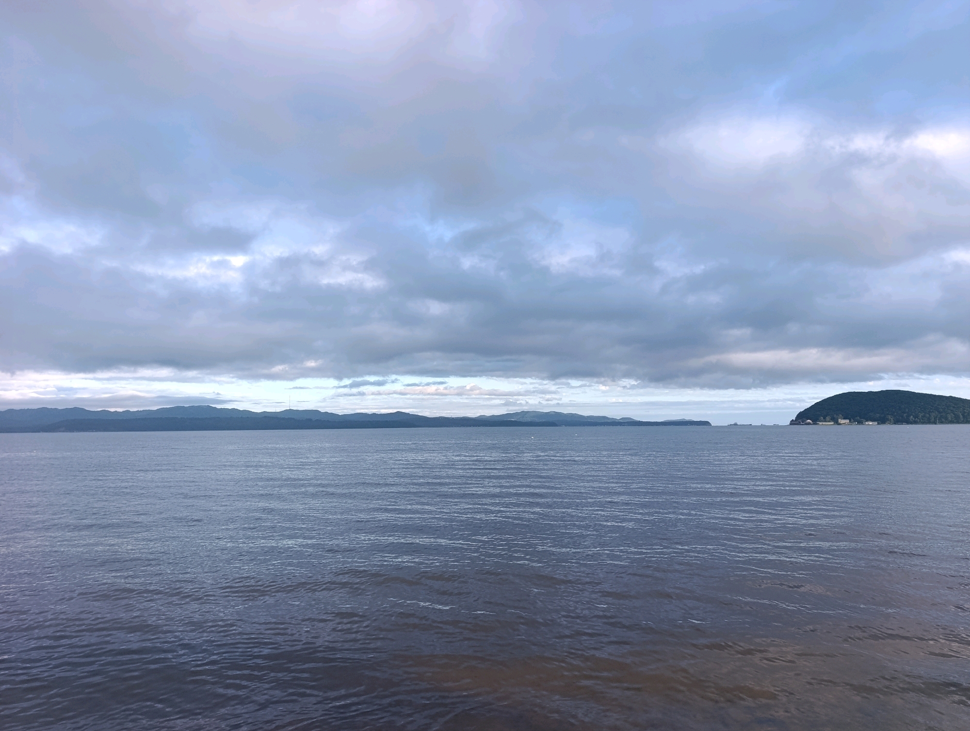 The image depicts a wide expanse of calm water under a cloudy sky. In the distance, a long, low mountain range stretches across the horizon. To the far right, a rounded, tree-covered island with buildings on its slope juts out into the water. The water itself is a dark, muted blue, with subtle ripples suggesting a gentle breeze. The sky is filled with layered clouds, ranging from light gray to darker, more ominous tones, with hints of brighter sky peeking through in places. The overall impression is one of a serene, somewhat overcast day at a coastal location. This scene is likely from Volchanec, Russia, given the characteristic coastal landscape and presence of islands.