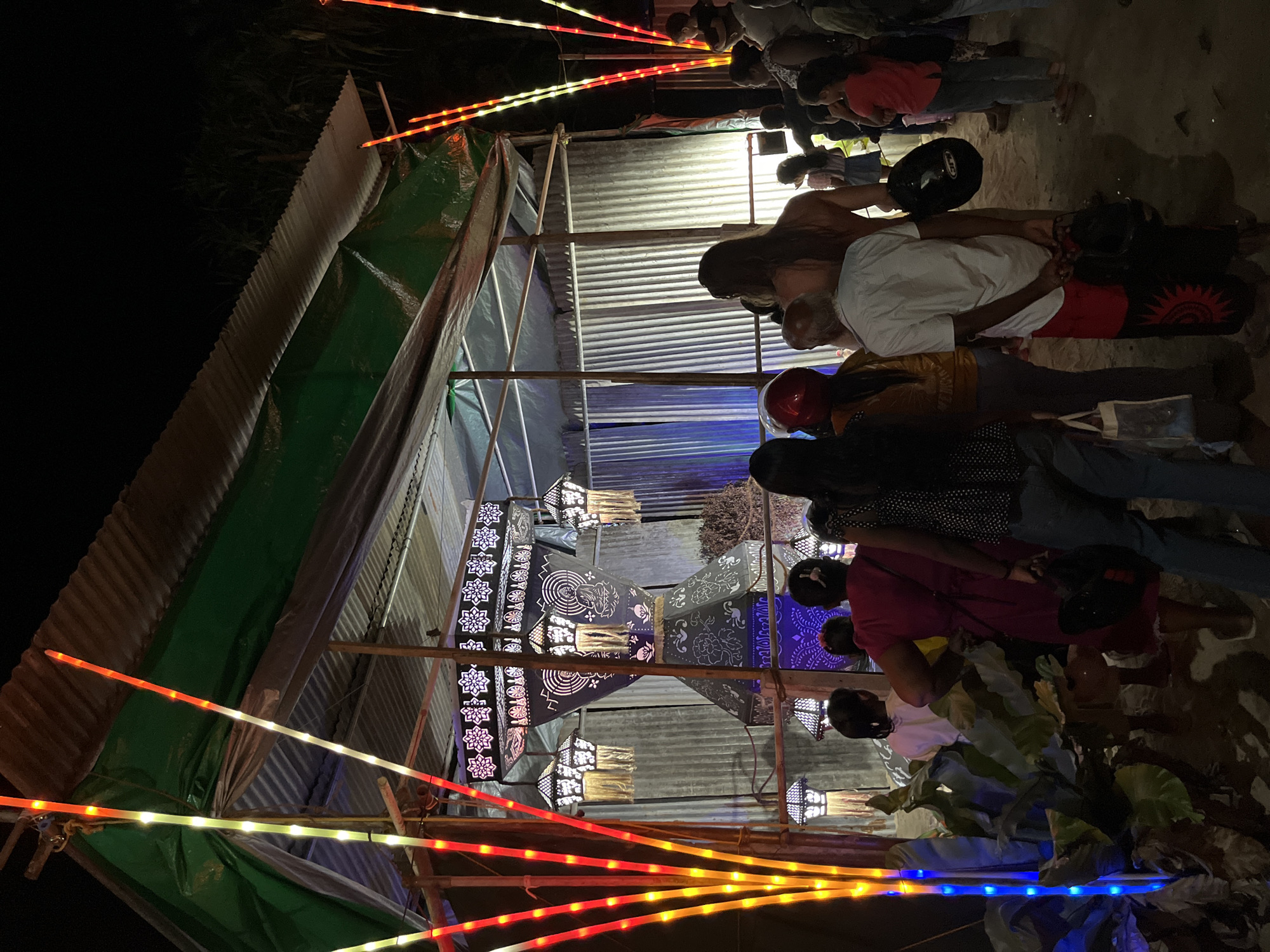 The image depicts an outdoor scene at night, likely in Habaraduwa, Sri Lanka. A group of people are gathered, observing or participating in an event centered around illuminated lanterns. The lanterns are intricately designed with decorative patterns, some appearing to be made of paper or fabric stretched over frames, and are lit from within, casting a warm glow. They hang from a makeshift structure supported by bamboo poles, which also appears to have corrugated metal roofing and green tarpaulin sides, suggesting a temporary setup, possibly for a festival or market stall.
Adding to the festive atmosphere, colorful LED string lights in red, yellow, and blue hues are strung along the foreground and sides of the structure, creating a vibrant and celebratory ambiance. The time of day is clearly night, evidenced by the darkness and the reliance on artificial lighting. The ground appears to be sandy or dusty. Some individuals are wearing helmets, possibly indicating they arrived on motorcycles. The overall mood seems to be one of community gathering and enjoyment, with the focus on the decorative lights and lanterns. No visible text is present in the image.