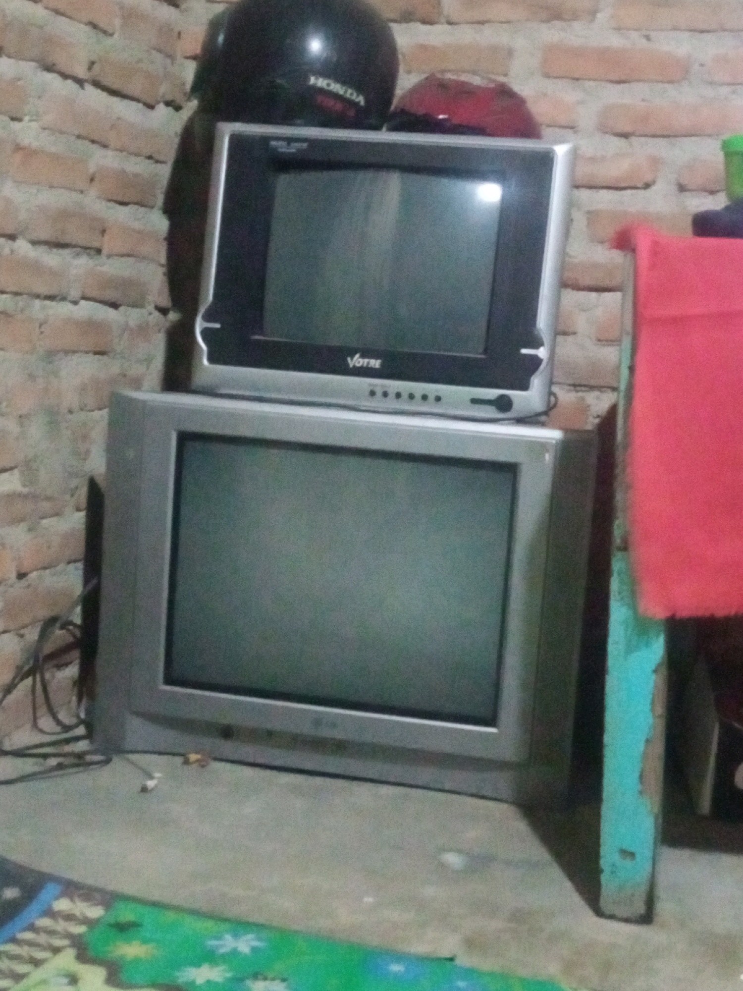 In a room with brick walls, two televisions are stacked on top of each other. The top television is smaller and silver, while the bottom television is larger and grey. A black motorcycle helmet with a "HONDA" logo is placed on top of the upper television. A red object, possibly another helmet, is also on top of the upper television. Various wires are visible near the bottom television, suggesting it is connected to power. A red cloth is draped over a blue and brown structure to the right of the televisions. The floor is covered in a colorful rug. The overall scene suggests a cluttered domestic environment.