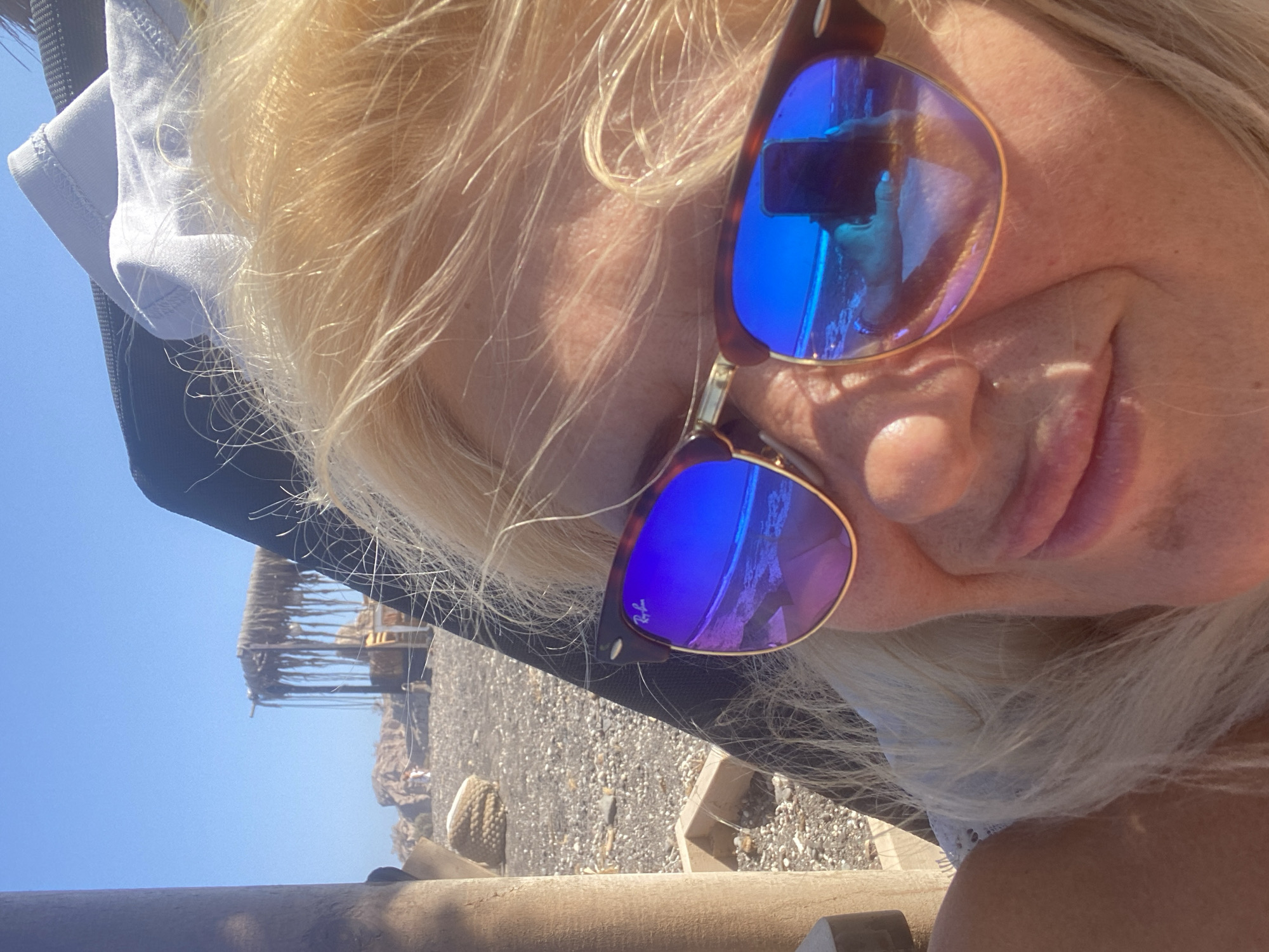 A blonde woman with fair skin wears sunglasses and poses for a selfie on a sunny beach. She is leaning back, with her face angled towards the camera. Her hair is light blonde and appears windblown. The sunglasses have blue mirrored lenses that reflect the sky, the sea, and the woman's face holding a phone. The background shows a dark, pebbly beach with some scattered rocks and a thatched-roof structure with hanging ropes. The sky is a clear, bright blue. The scene suggests a vacation or leisure setting. The location is the Thira Municipal Unit, Greece.